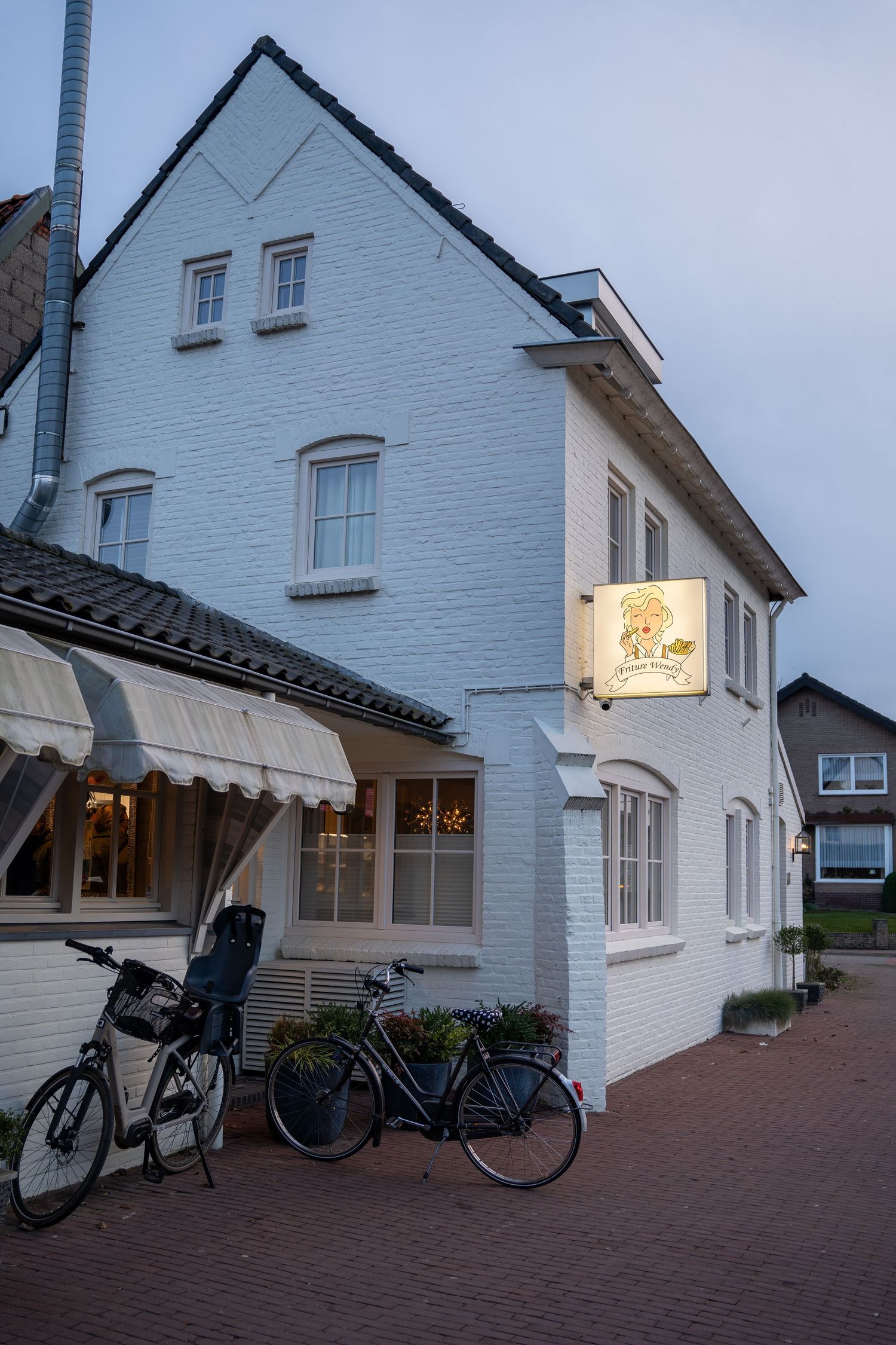 White brick Dutch restaurant building at dusk with bicycles parked outside and illuminated sign.
