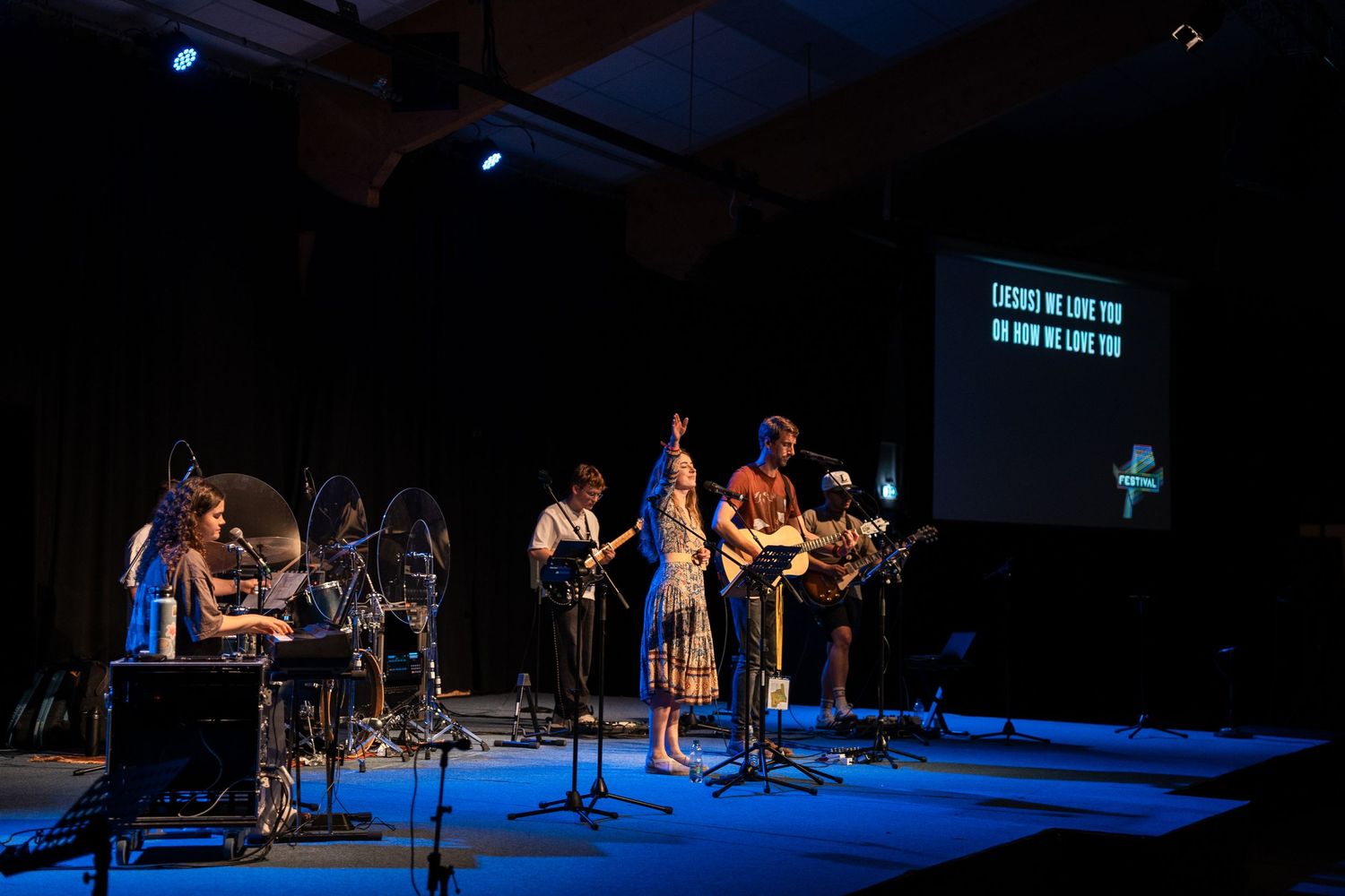 Live worship band performing on a blue-lit stage with lyrics projected on a screen.