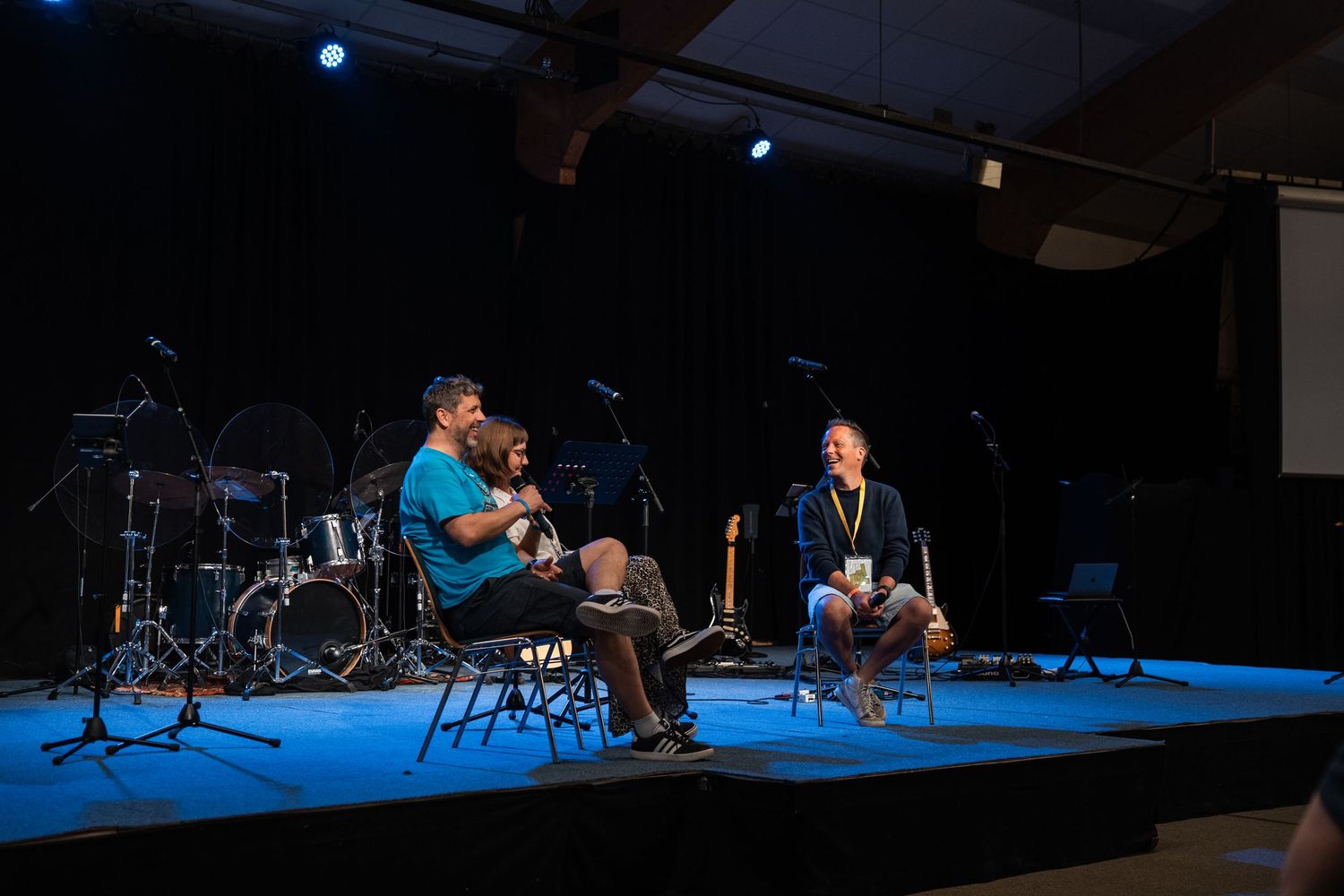 Three people sit on stage chairs in conversation during a Christian conference panel discussion with drums behind.