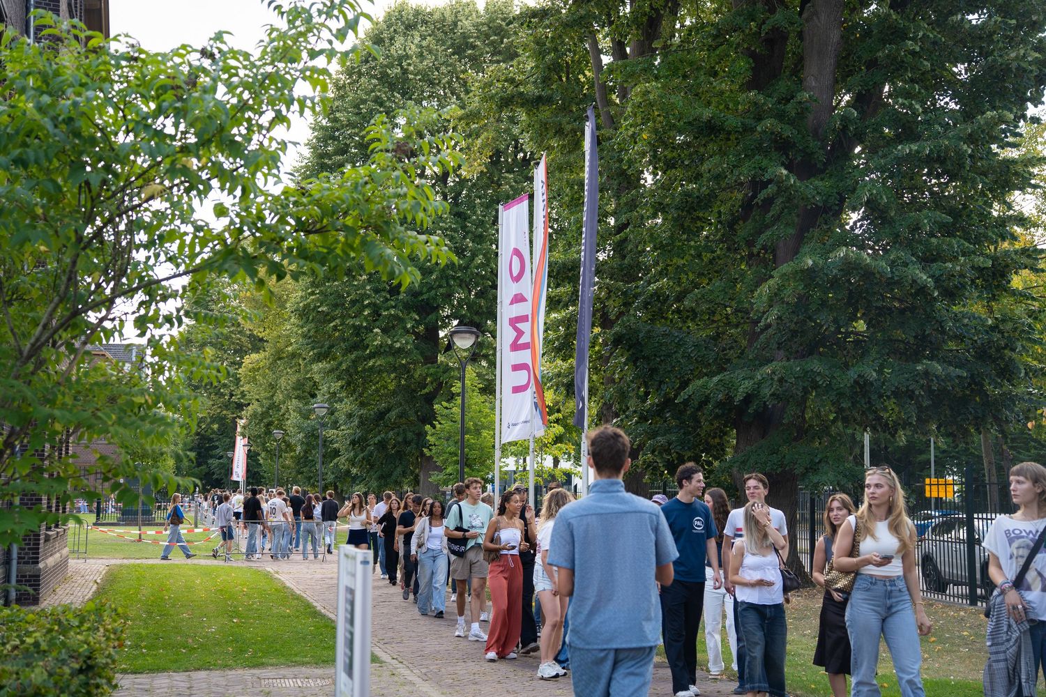 Students walking along tree-lined campus path with INKOM event banners displayed.