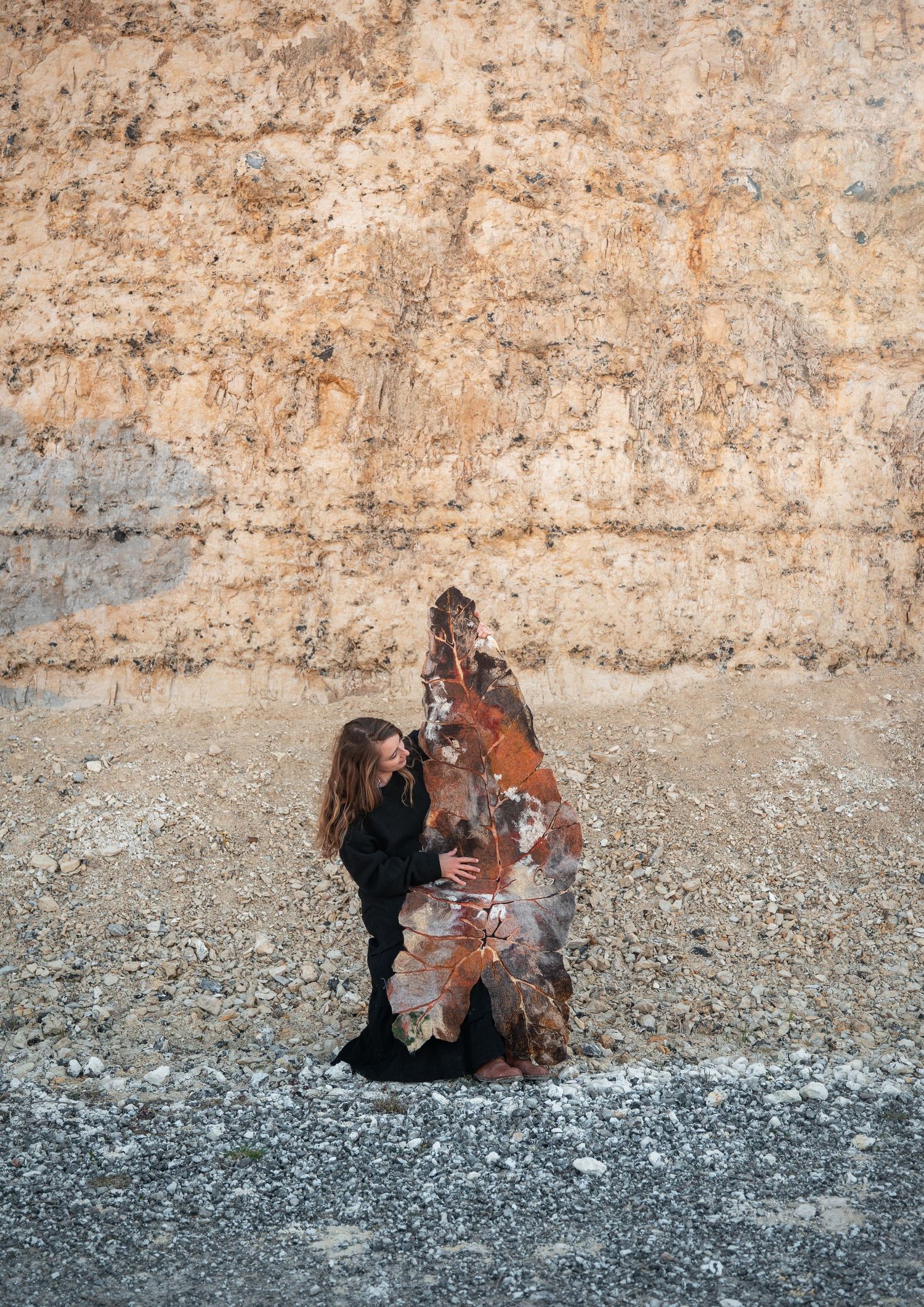 Woman in black interacting with a large rust-colored felted sculpture against a rocky cliff wall.