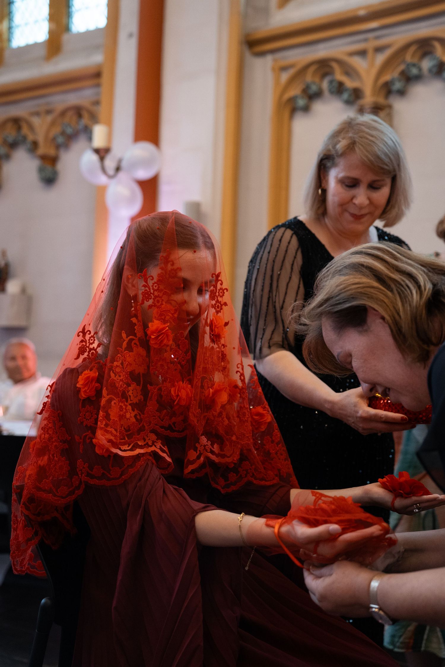 Bride in a red lace gown and veil is assisted by two women during a wedding ceremony indoors.