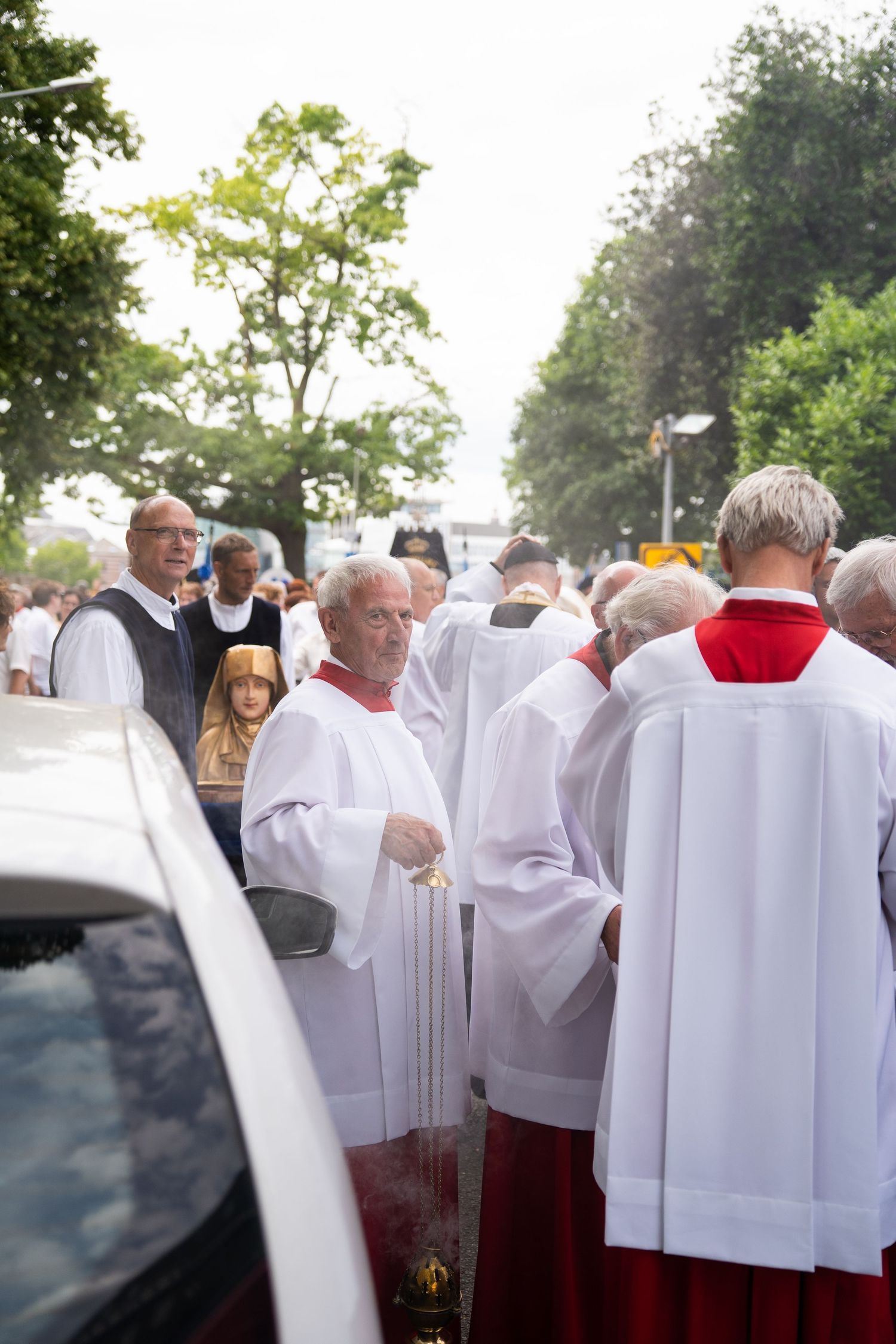 Clergy in white and red robes participate in a religious street procession carrying sacred items and incense.