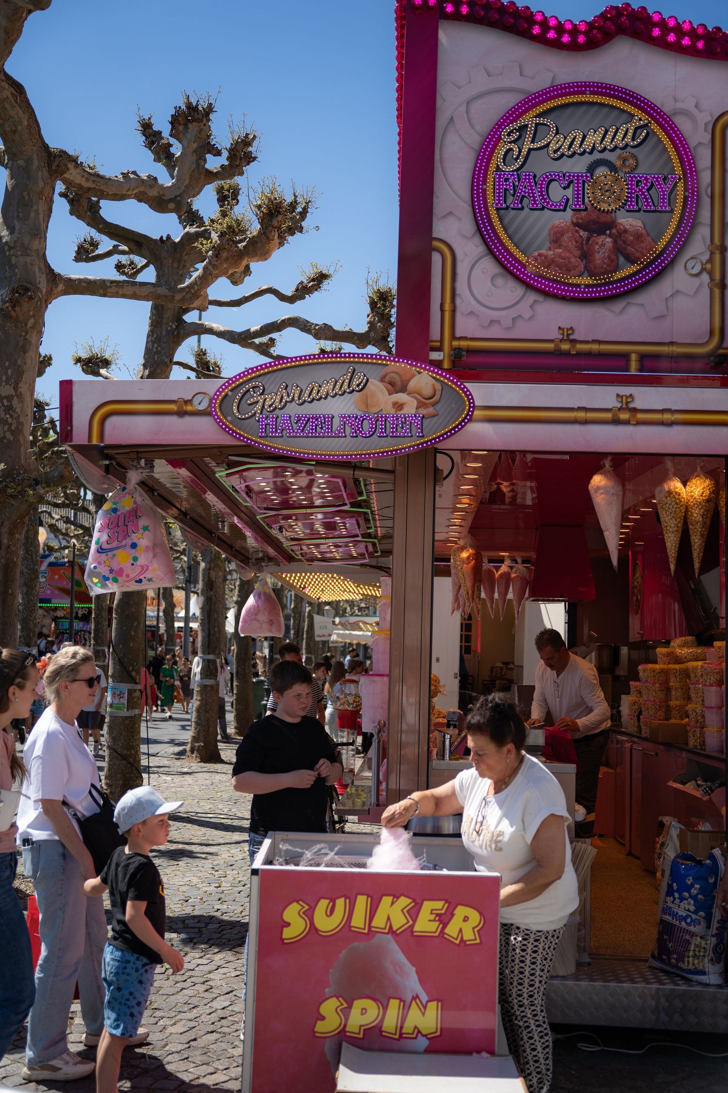 Vendor sells cotton candy at a Peanut Factory food stall during a busy outdoor fair on a sunny day.