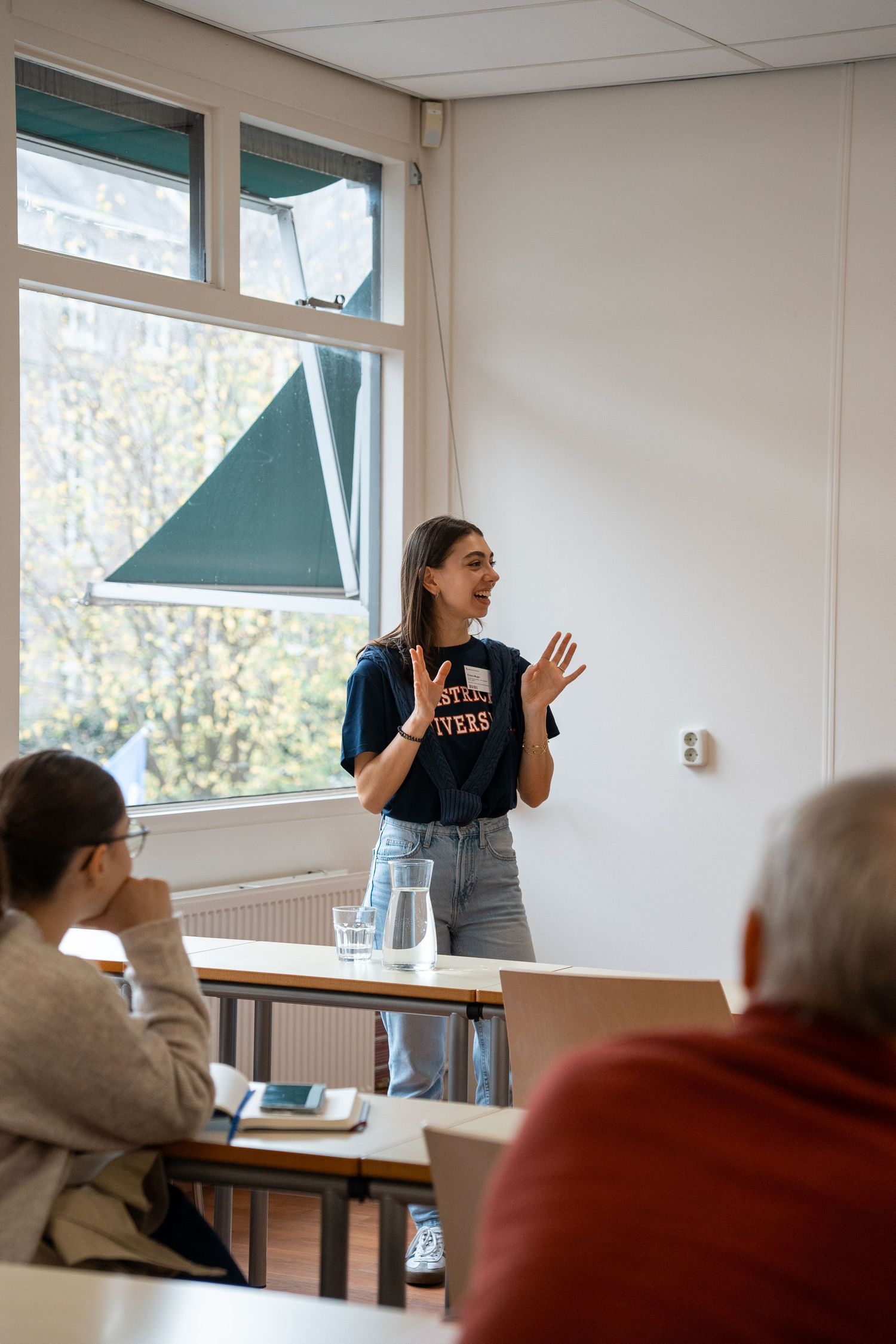 A young female student presents to classmates in a sunlit university classroom while gesturing expressively.