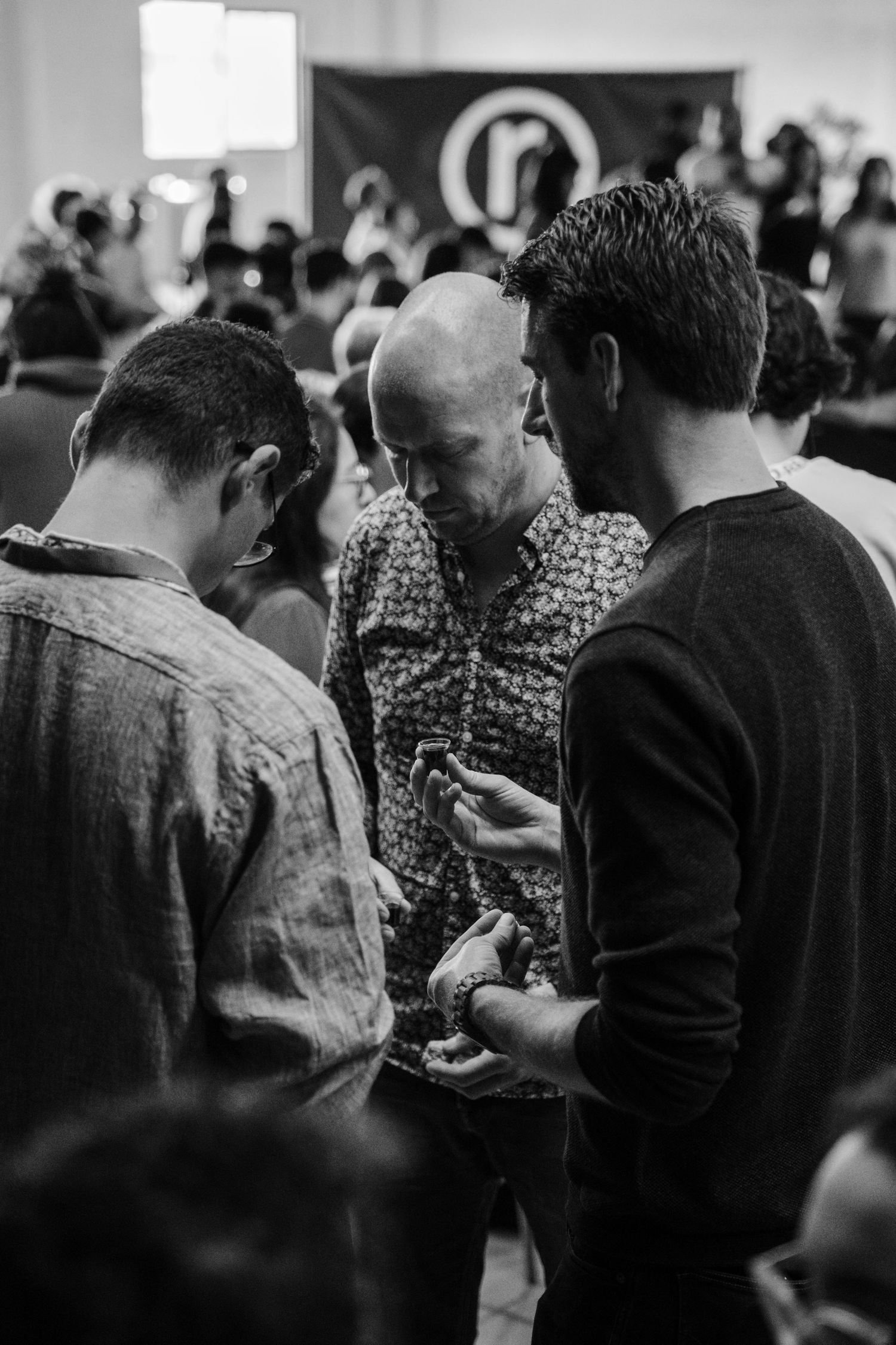 Three men in conversation at black and white church gathering with r. logo banner background.