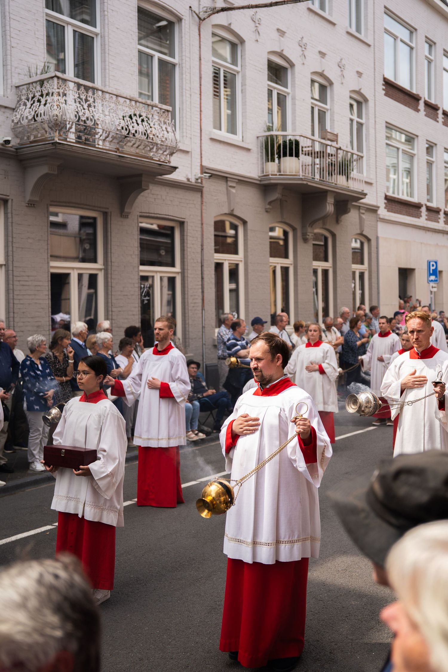 Altar servers in white and red robes carry incense thuribles during a Catholic street procession.