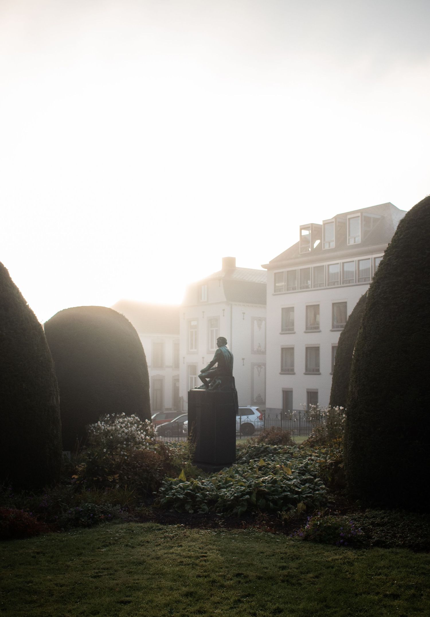 Bronze statue on a pedestal surrounded by sculpted topiary hedges in a misty garden at sunrise.