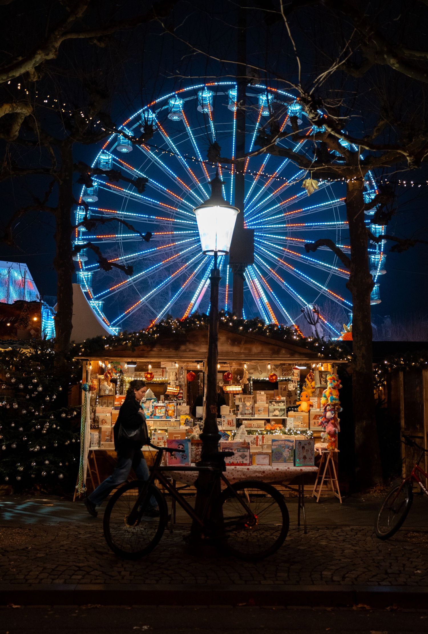 Glowing Ferris wheel behind a Christmas market stall with a bicycle parked in front at night.