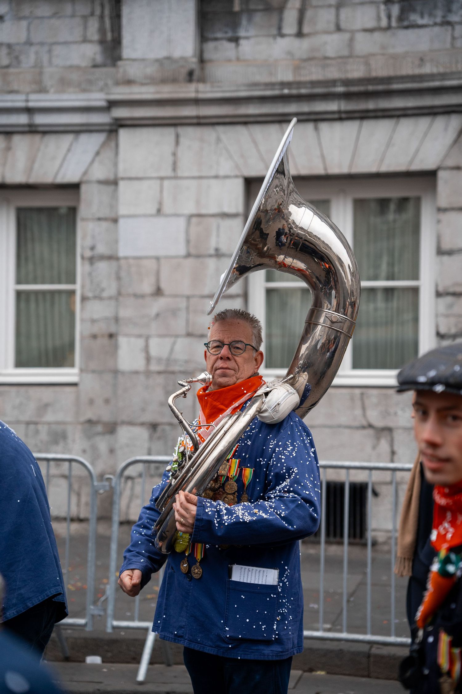 Musician playing a sousaphone in a parade, wearing a blue sequined jacket with medals and an orange scarf.