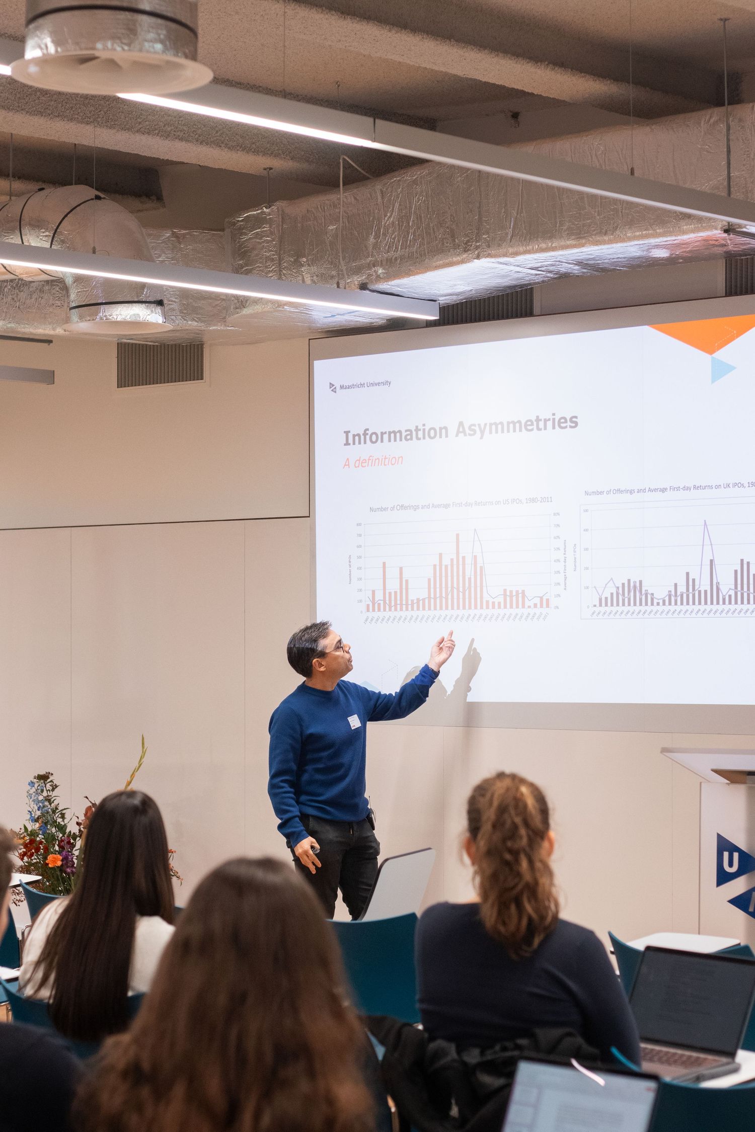 A professor presents information asymmetries data on a large screen to students in a university lecture.