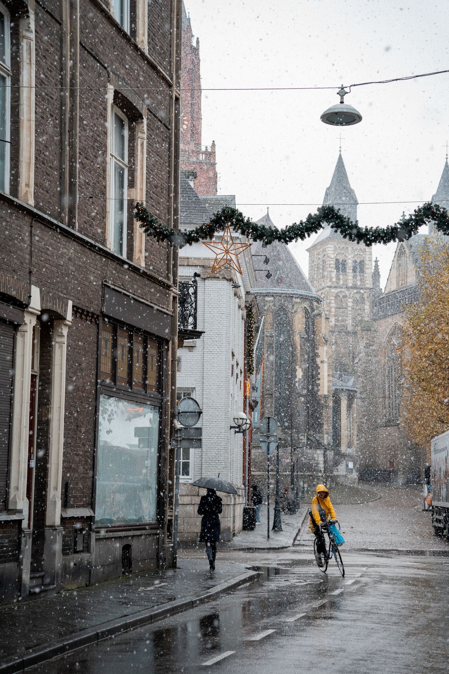 Cyclists and pedestrians on a rainy snowy day in a historic European street with Christmas decorations.