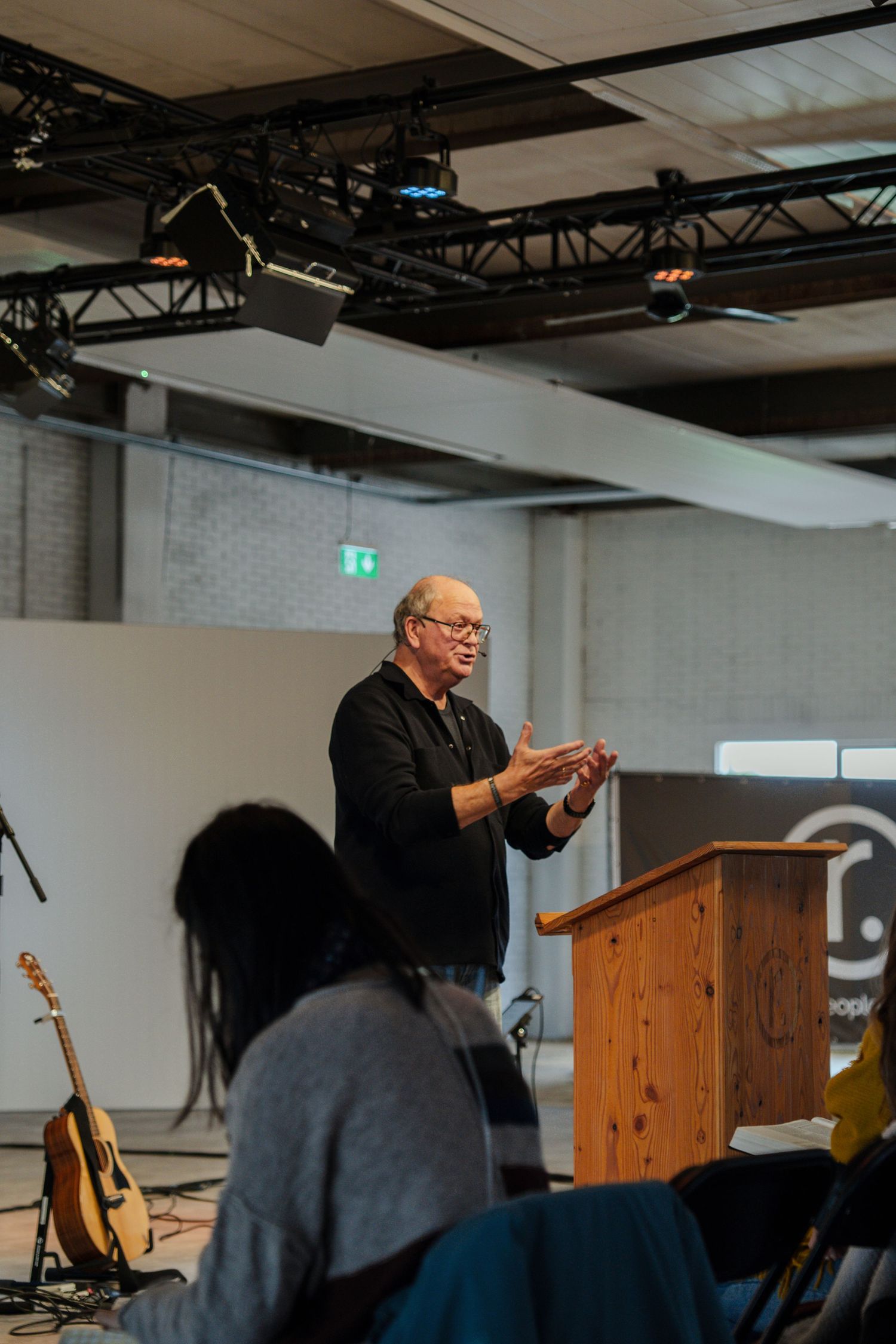 Older man in black giving a speech at a wooden podium in a modern event space with stage lighting.
