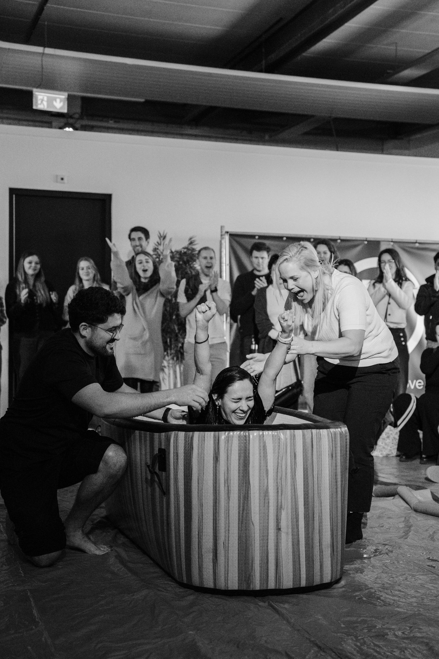 Black and white photo of a joyful water baptism ceremony with supporters cheering in an indoor venue.