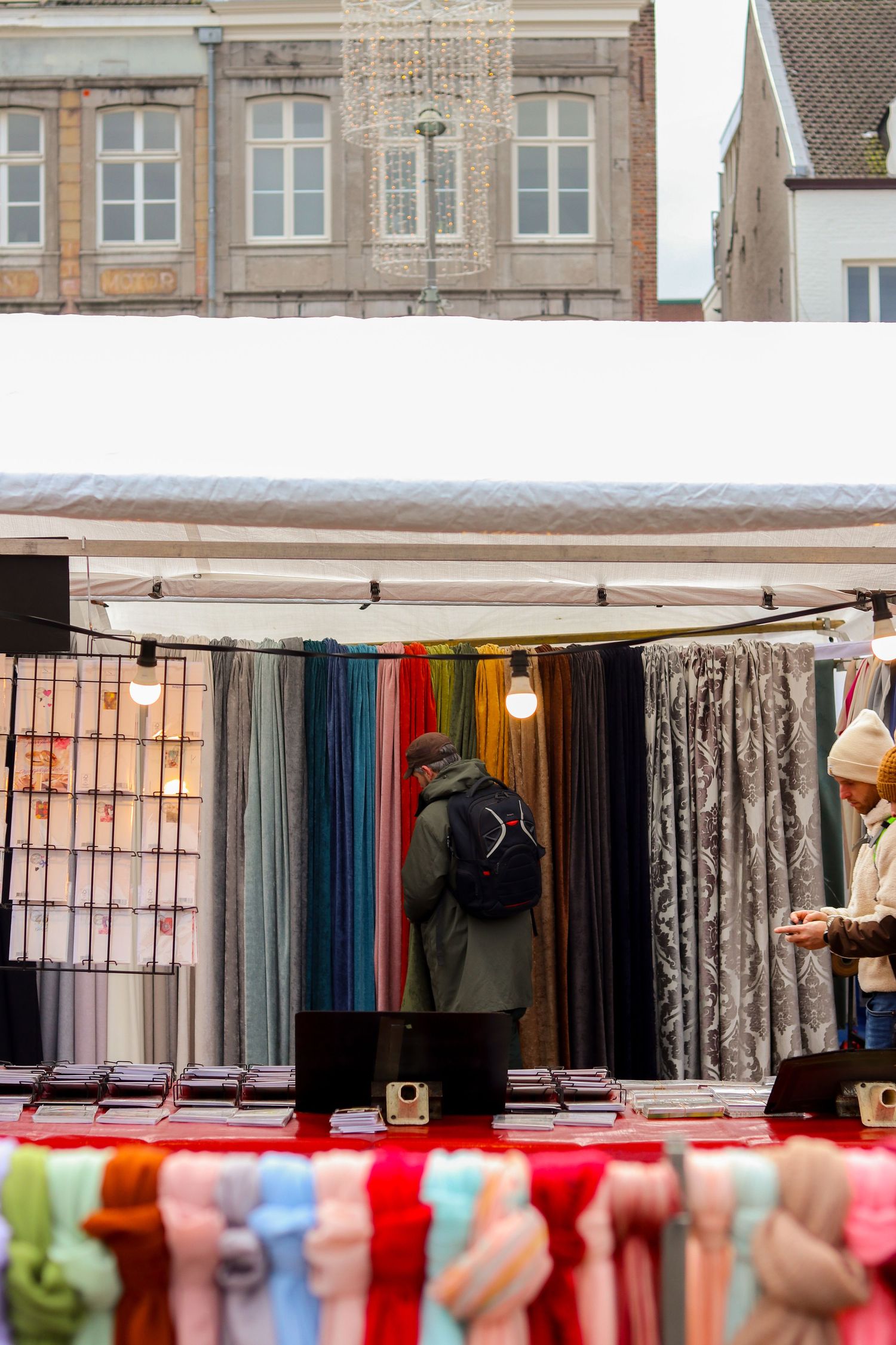 Colorful fabric and scarf market stall with shoppers browsing textiles outdoors.