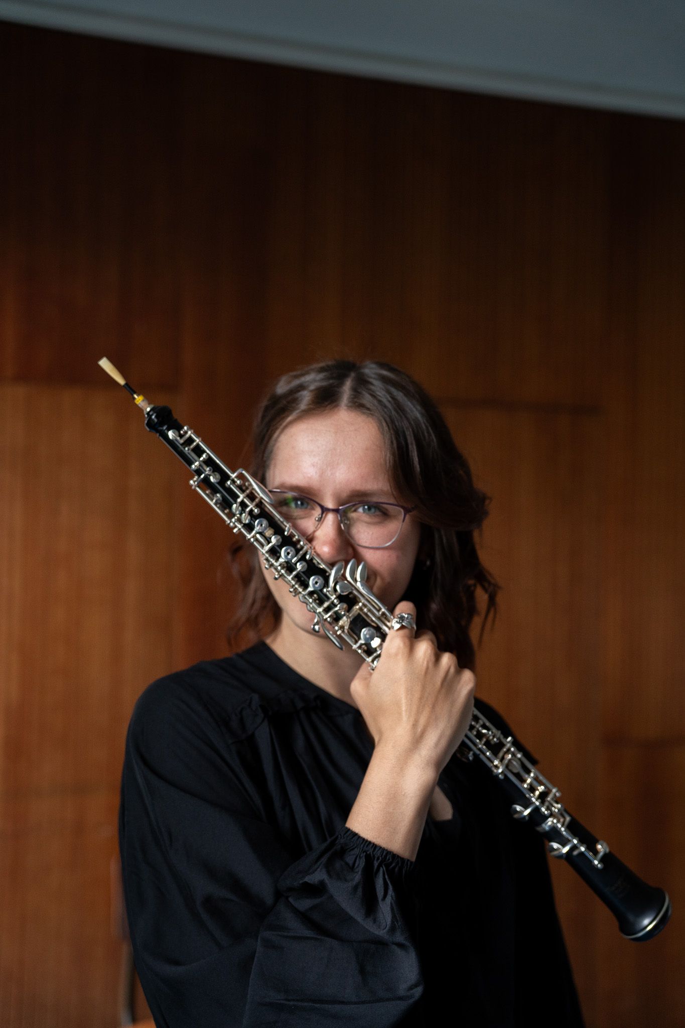 Female oboist in black outfit holding her oboe up near her face against a wooden panel background.