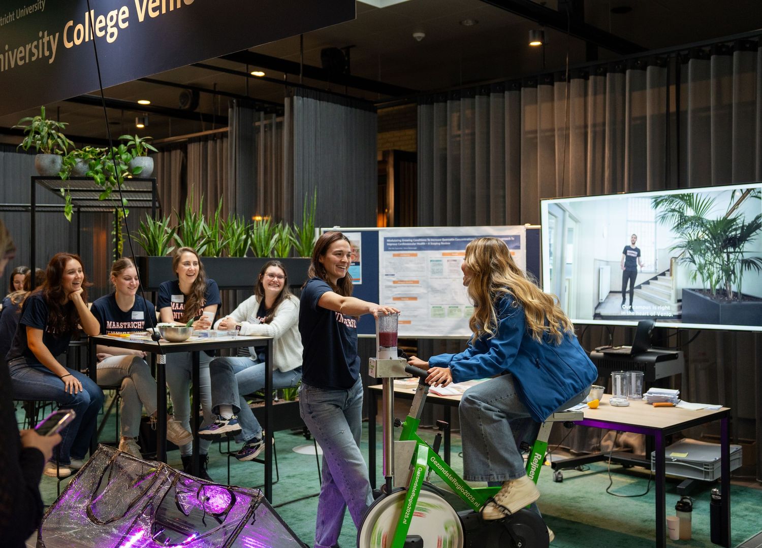 Lively University College Venlo exhibition booth with students engaging around a stationary green bicycle display.