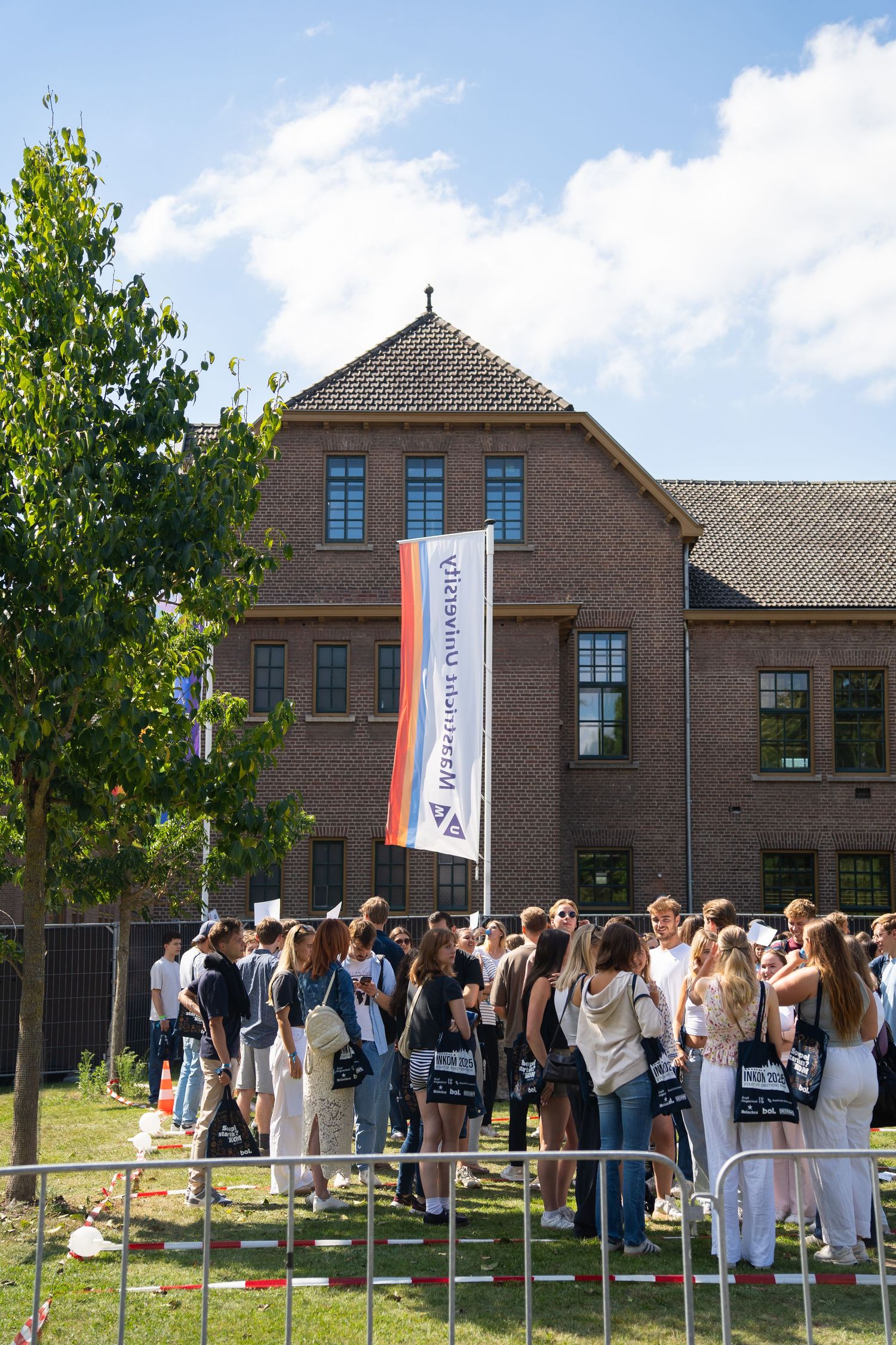 Students gather outside a brick university building beneath a Maastricht University flag on a sunny day.