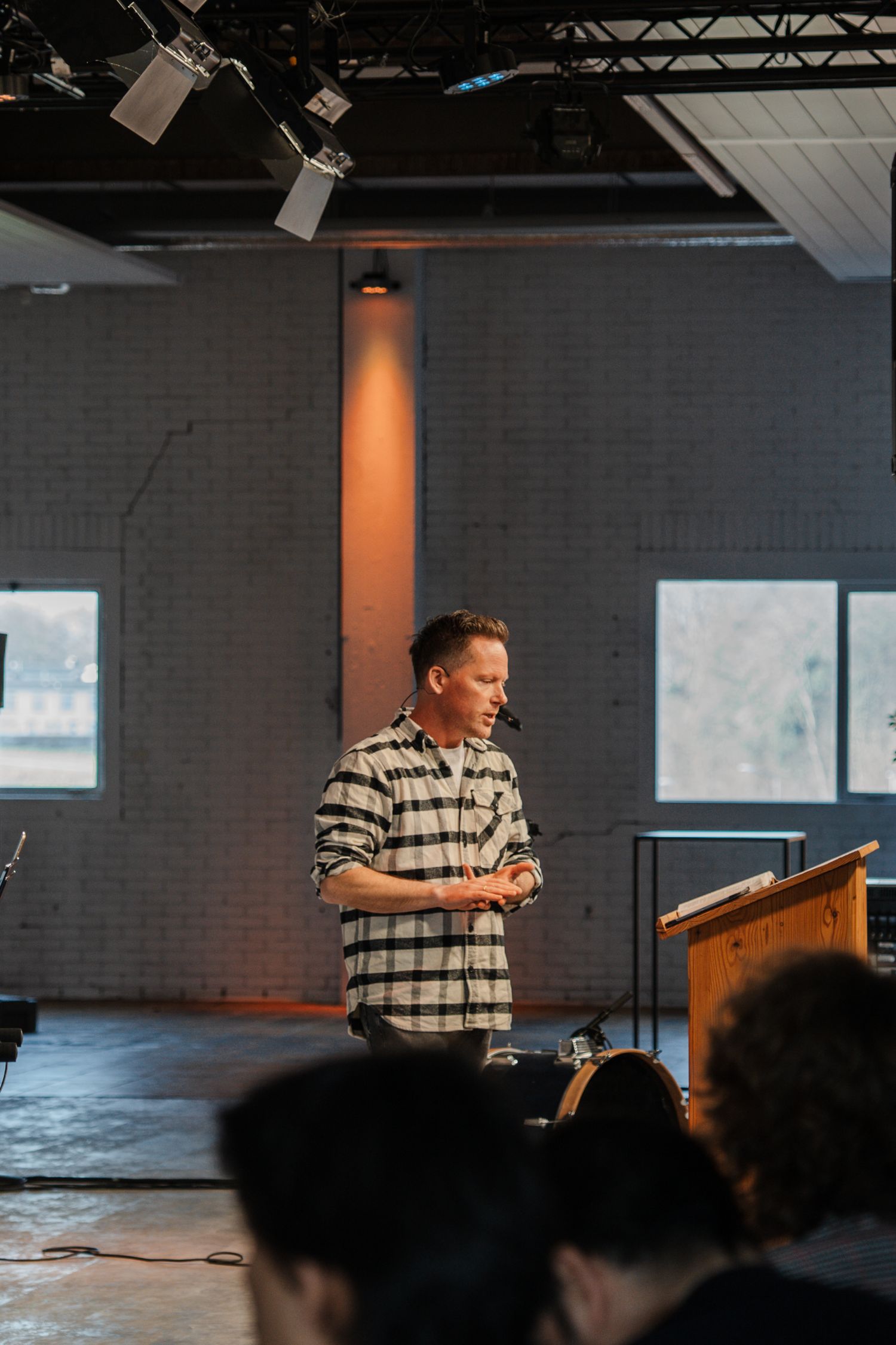 Male pastor in plaid shirt delivering a sermon on a modern church stage with brick wall backdrop.