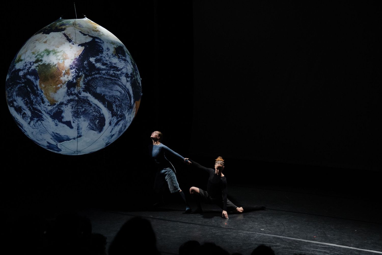Two dancers performing on dark stage with large suspended Earth globe in theatrical dance show.