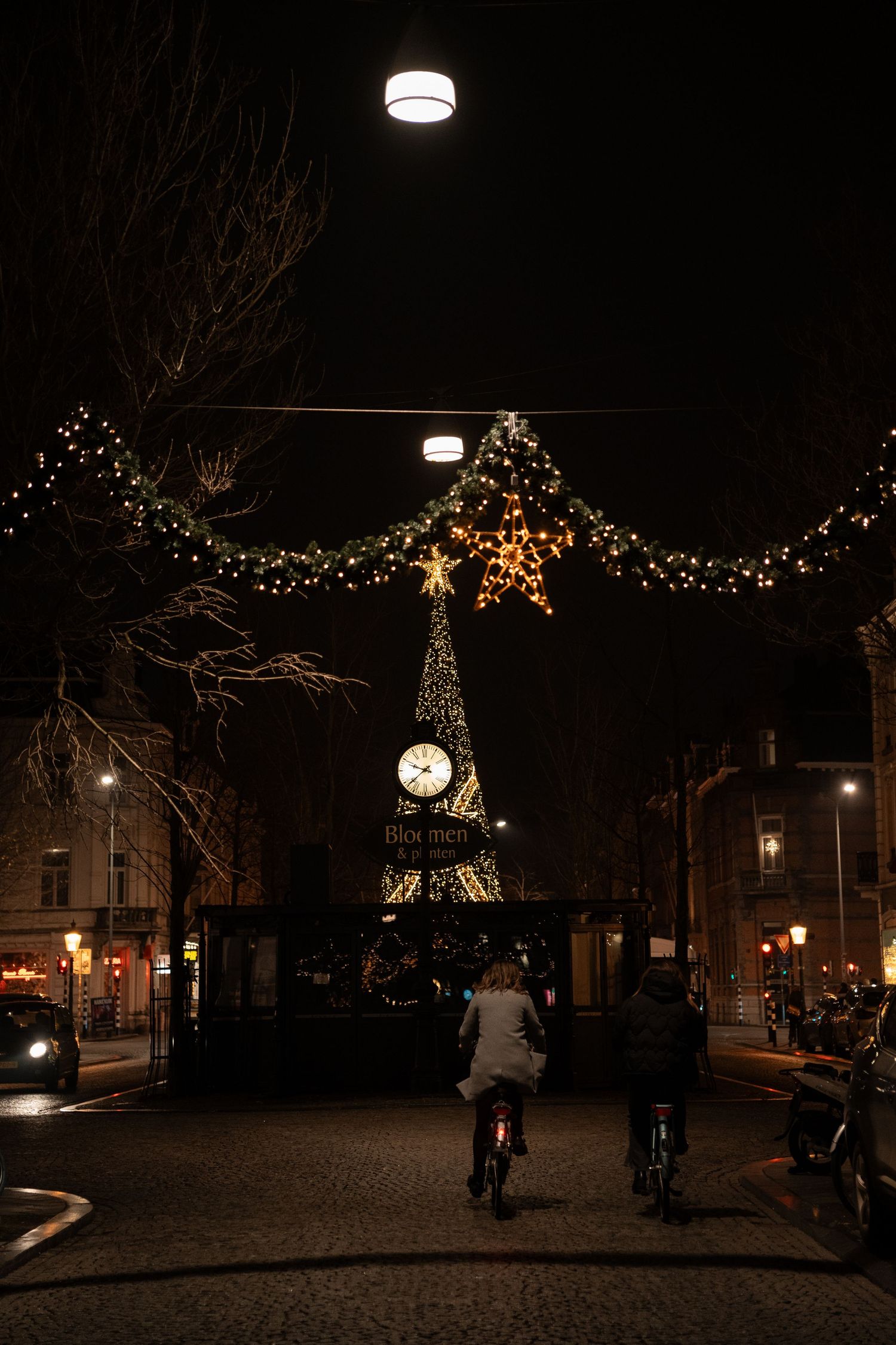 Cyclists riding at night past a glowing Christmas tree and clock adorned with festive lights in a city.