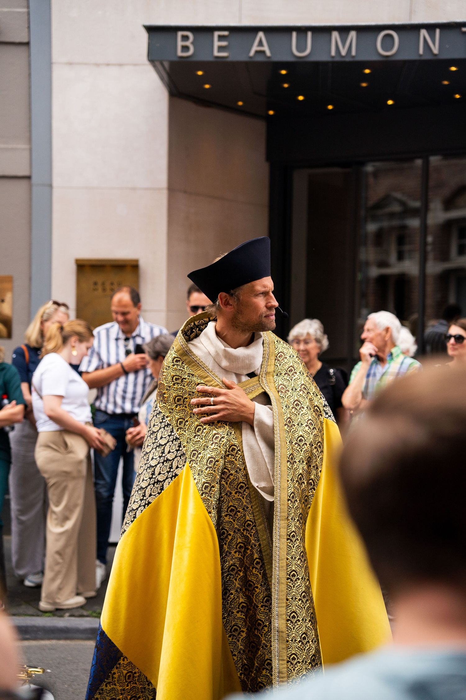 Catholic priest in golden vestments and biretta hat leads a religious procession through city streets.