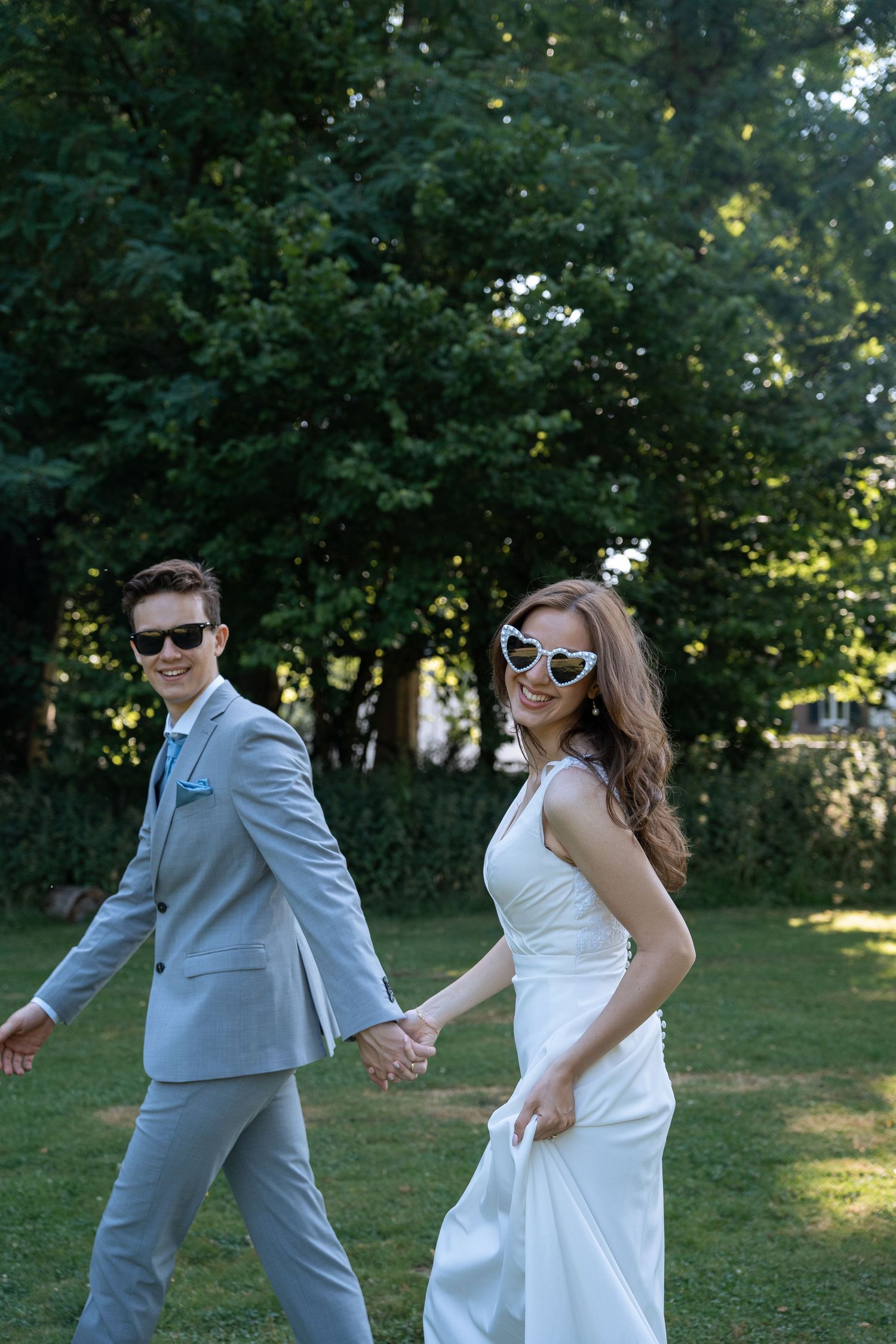 Smiling bride and groom holding hands walking in a park wearing stylish sunglasses.