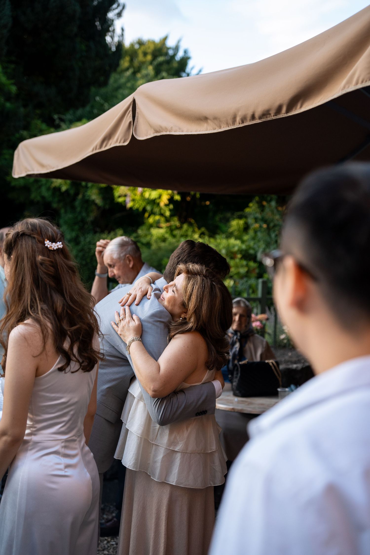 Guests hug and celebrate at an outdoor wedding reception surrounded by lush green trees and warm sunlight.