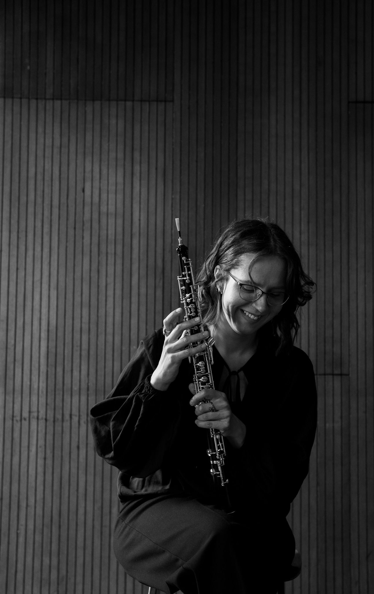 Black and white portrait of smiling woman holding oboe instrument against striped background.