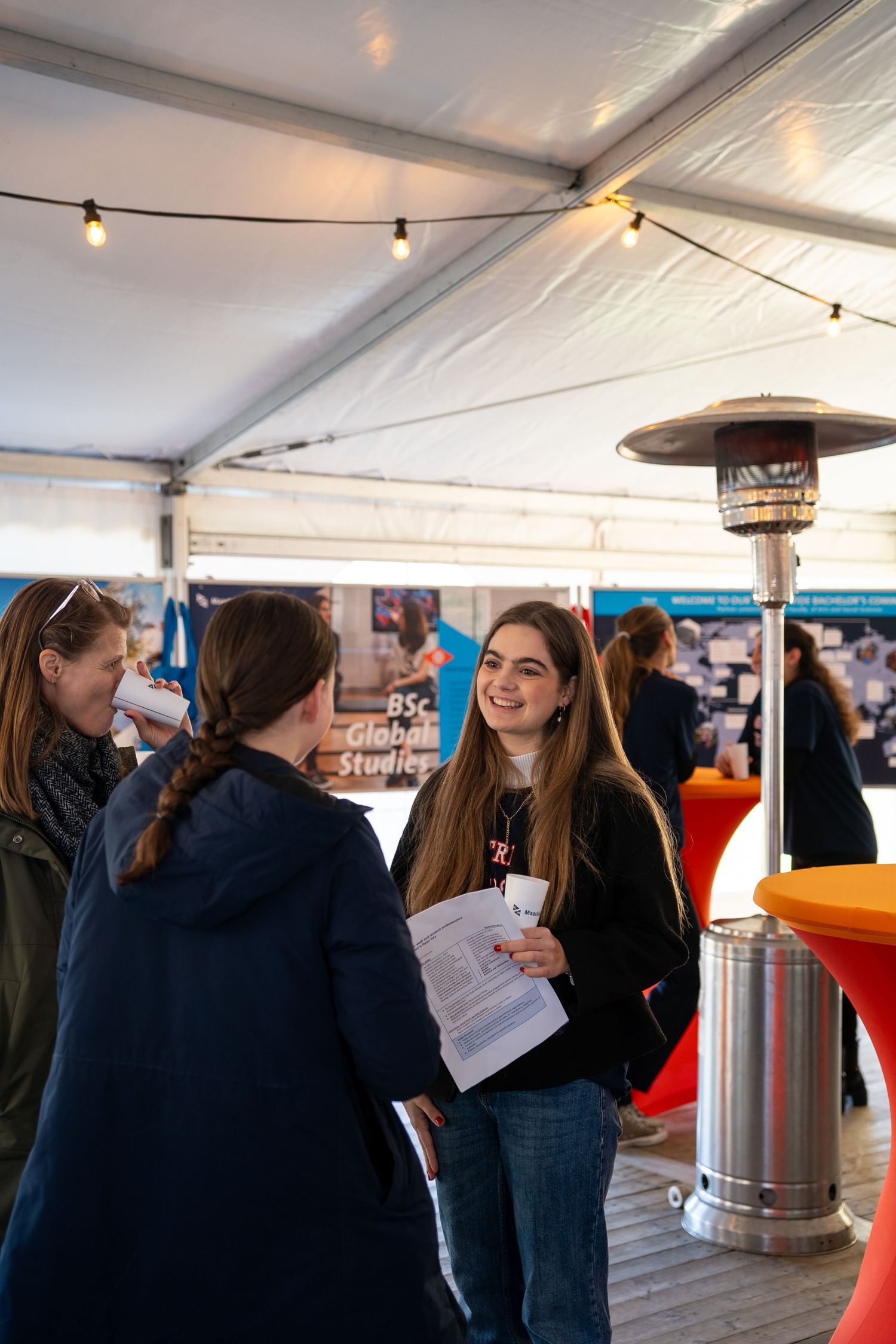 Two smiling young women chat near a patio heater inside a tent at a university open day fair.