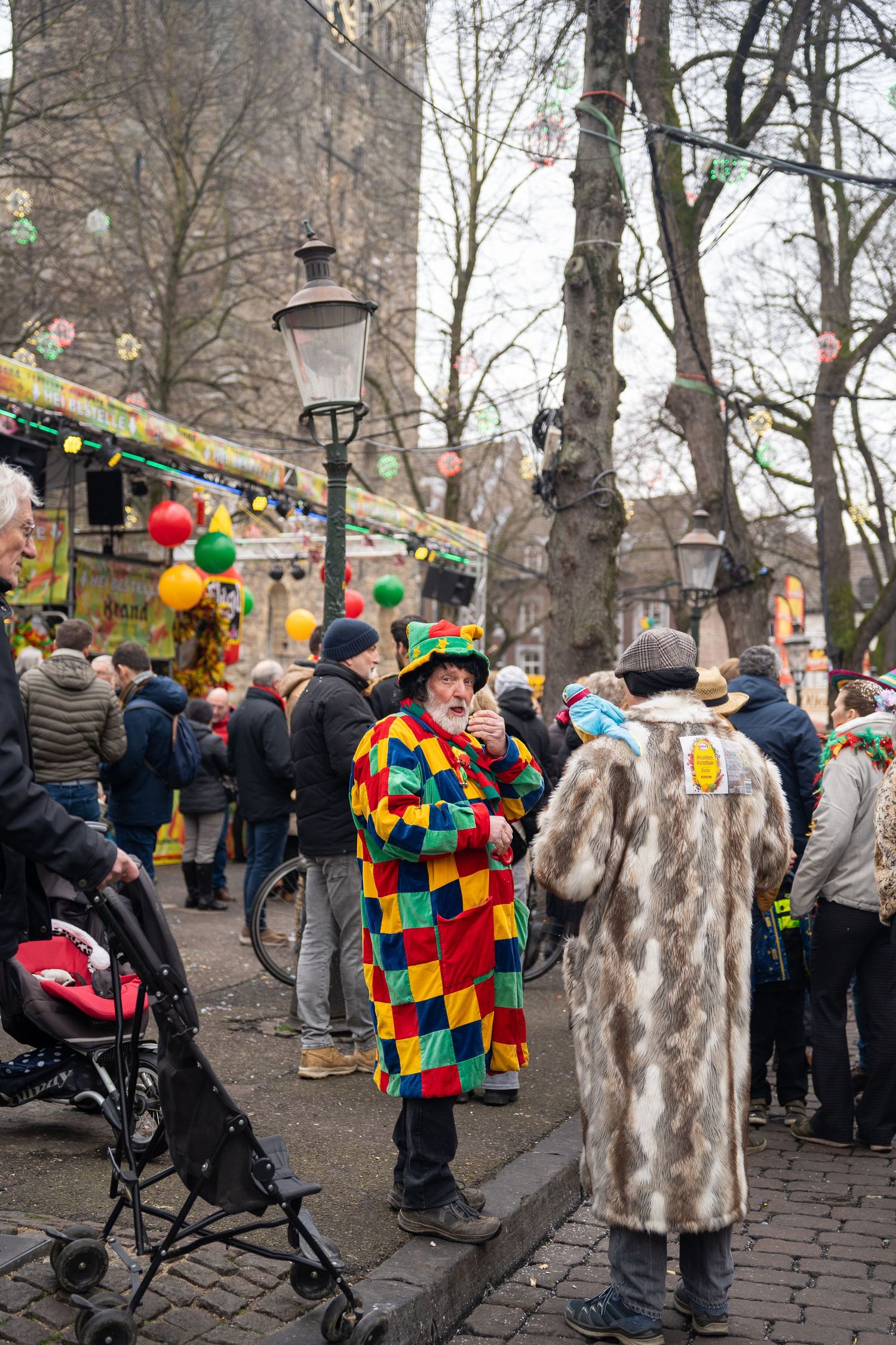 Person in colorful harlequin costume at a festive outdoor carnival market event.