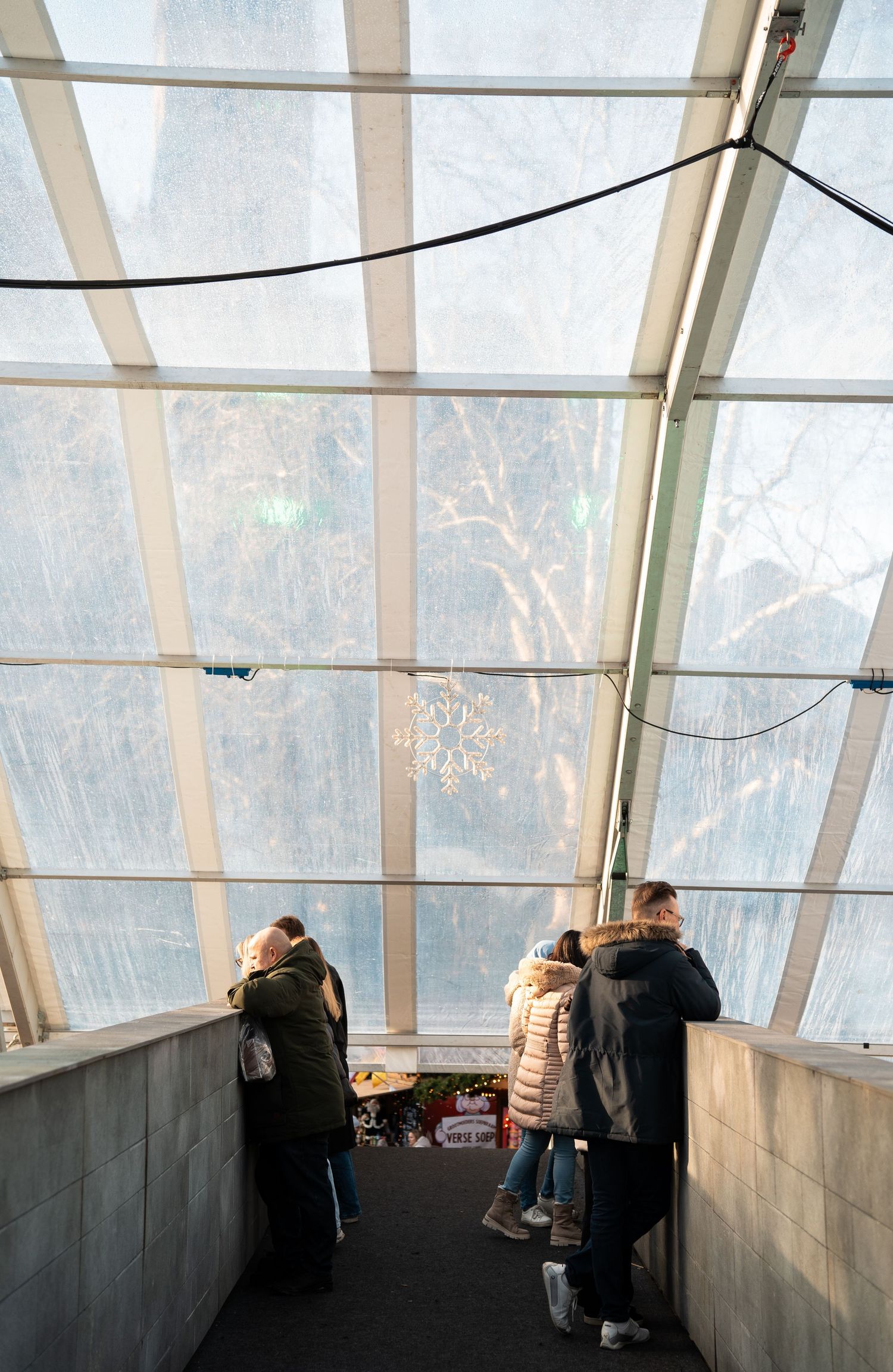 Visitors looking over a railing inside a glass-roofed structure at a winter Christmas market.