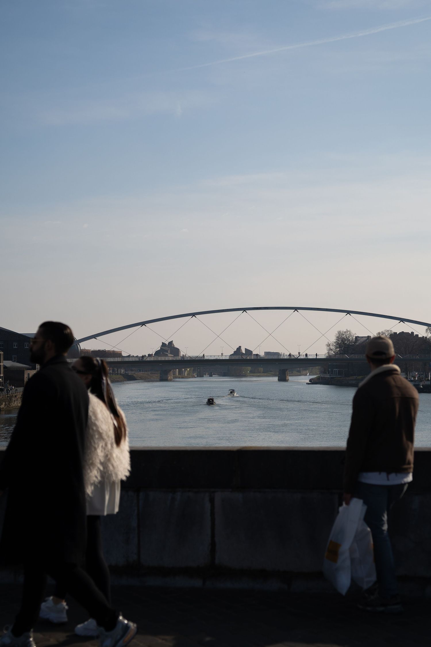 People walking along a riverside promenade with an arched pedestrian bridge spanning the river in the background.