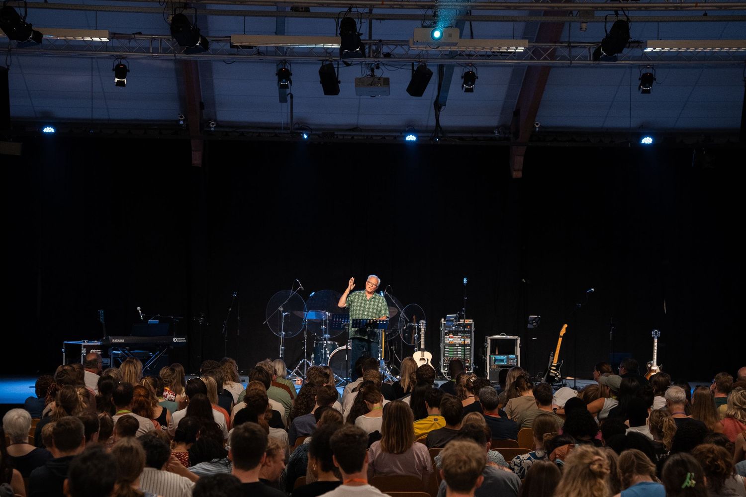 Speaker presenting on a dark stage to a large seated audience at an indoor conference event.