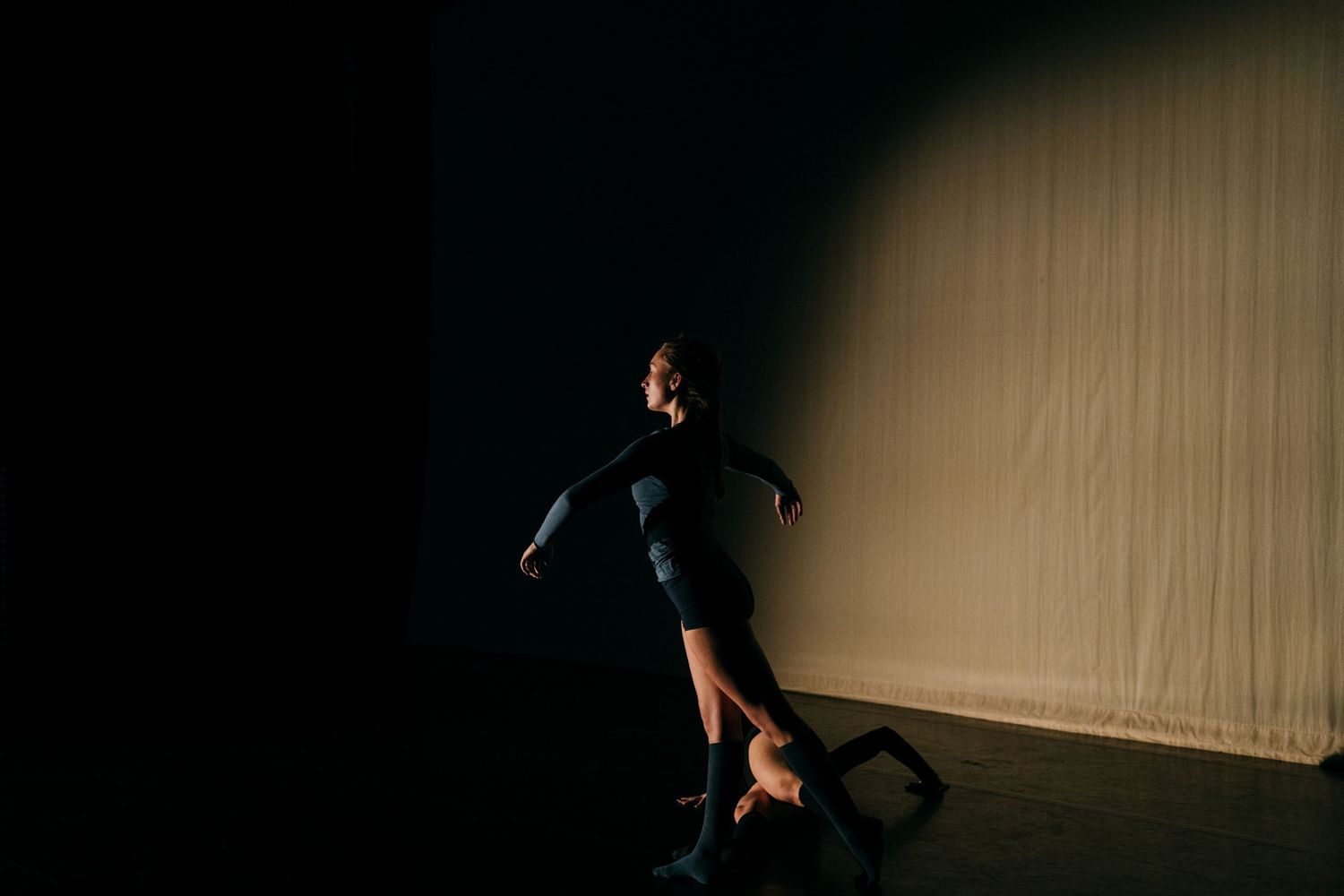 Dancer in black outfit performs near white backdrop in dramatic low-light contemporary dance piece.