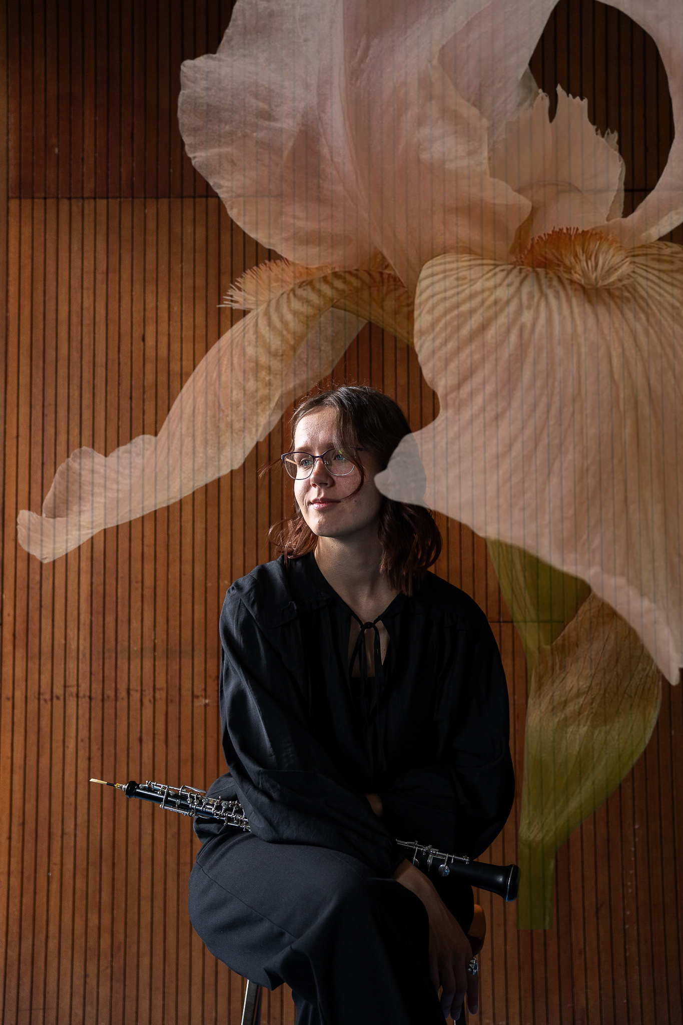 Female oboist sitting with clarinet before projected iris flower on wooden panel background.