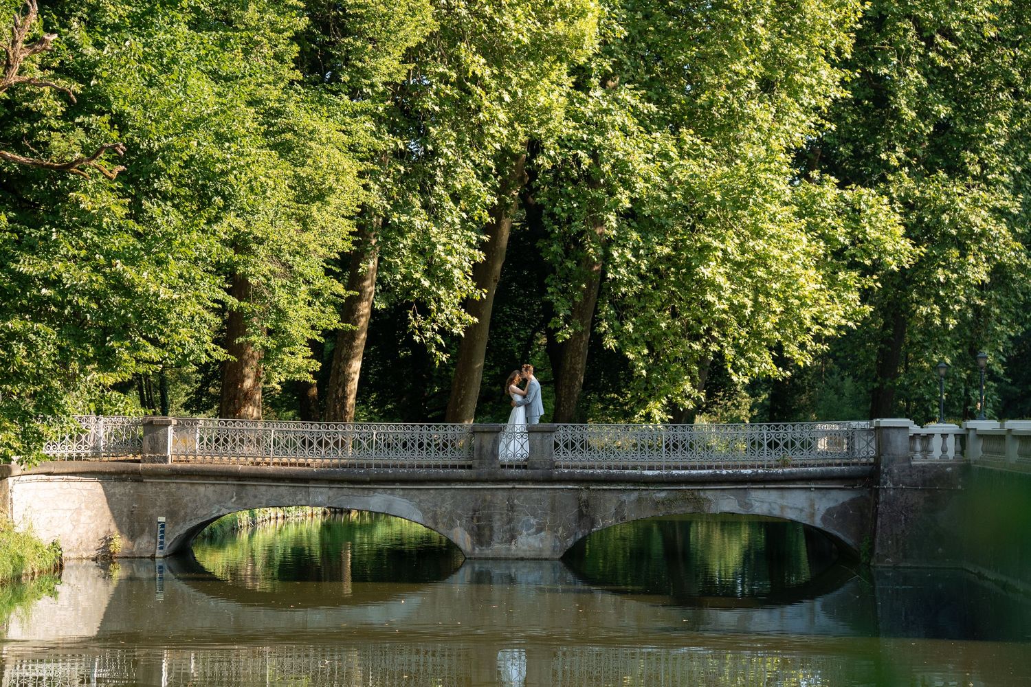Couple embraces on an ornate stone bridge surrounded by lush green trees reflected in calm water below.