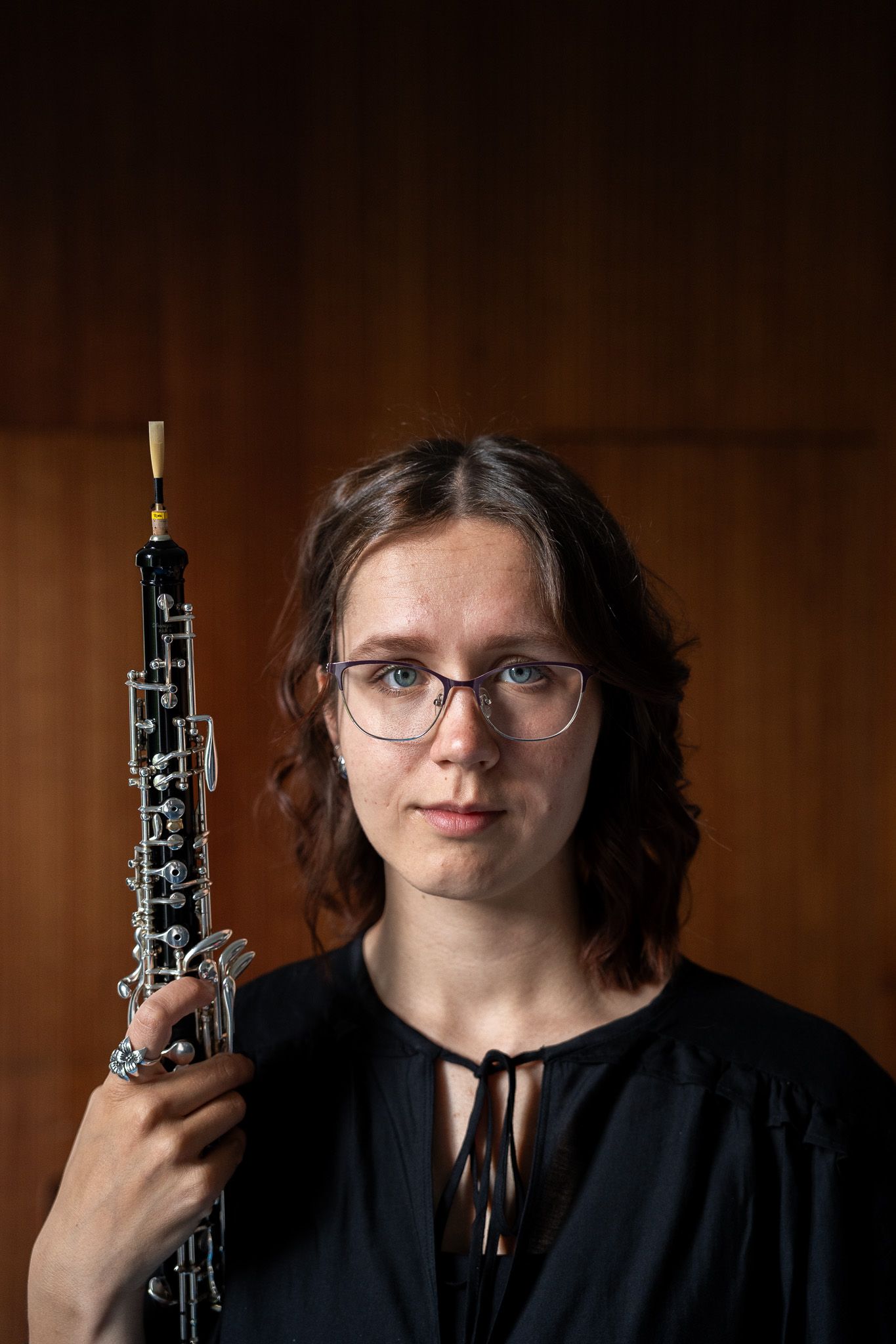 Close-up portrait of a female oboist holding her oboe upright beside her face against a wooden background.