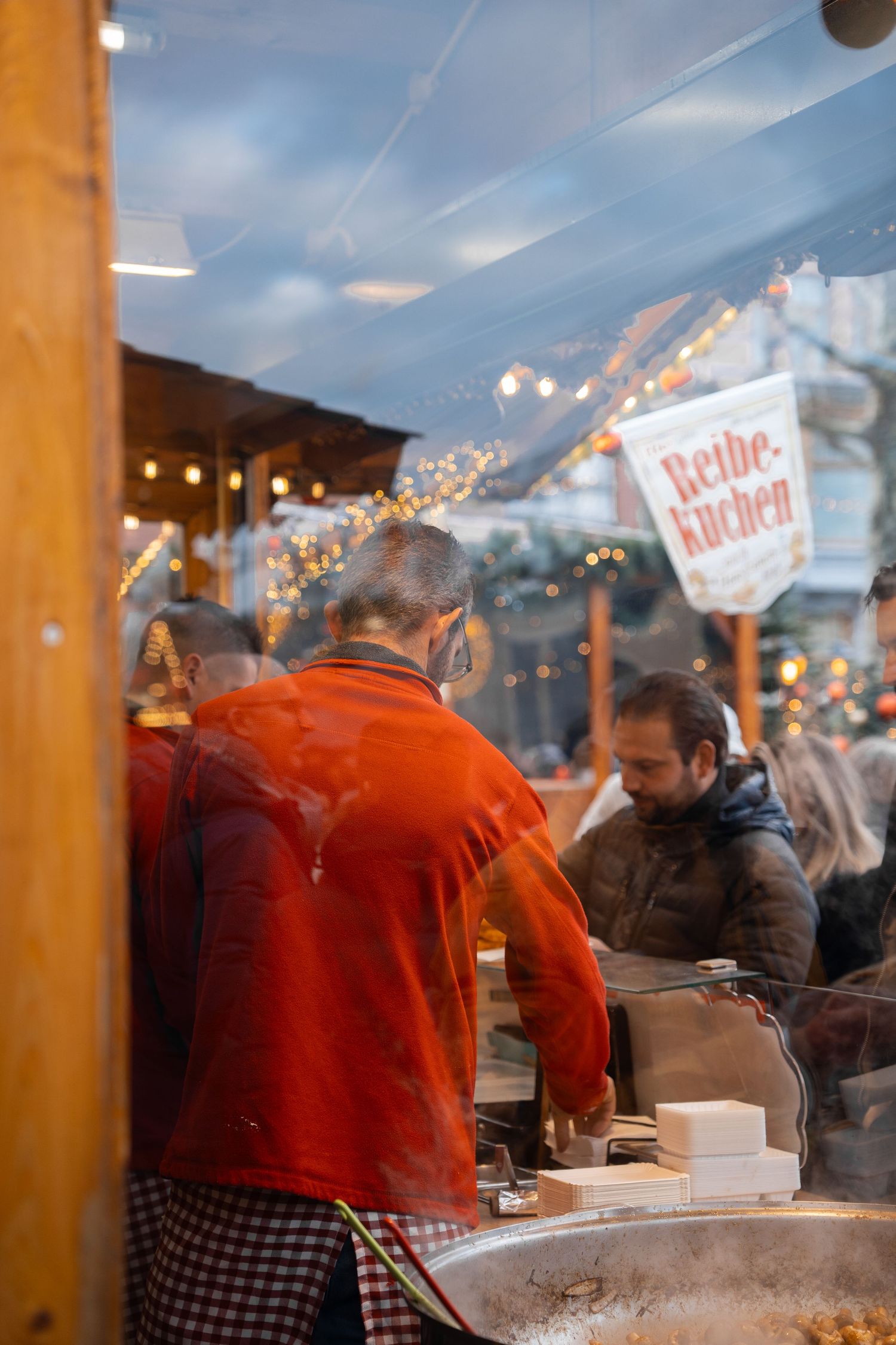 Vendor in red jacket serving customers at a Reibekuchen food stall at a festive Christmas market.
