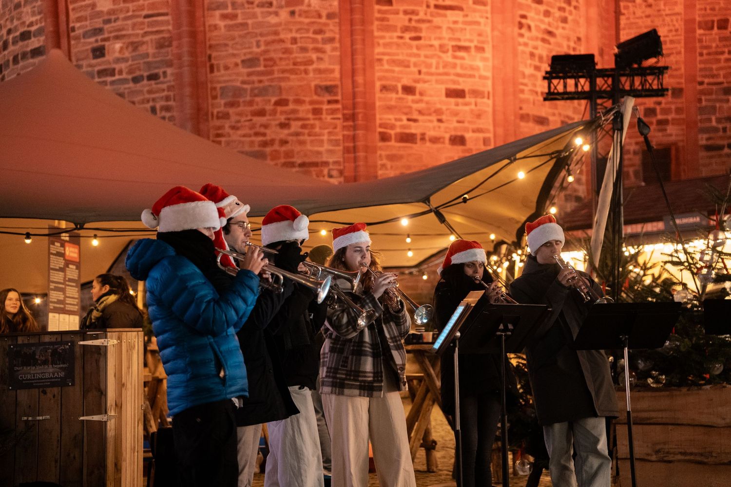 Musicians wearing Santa hats performing with brass instruments at an outdoor Christmas market at night.