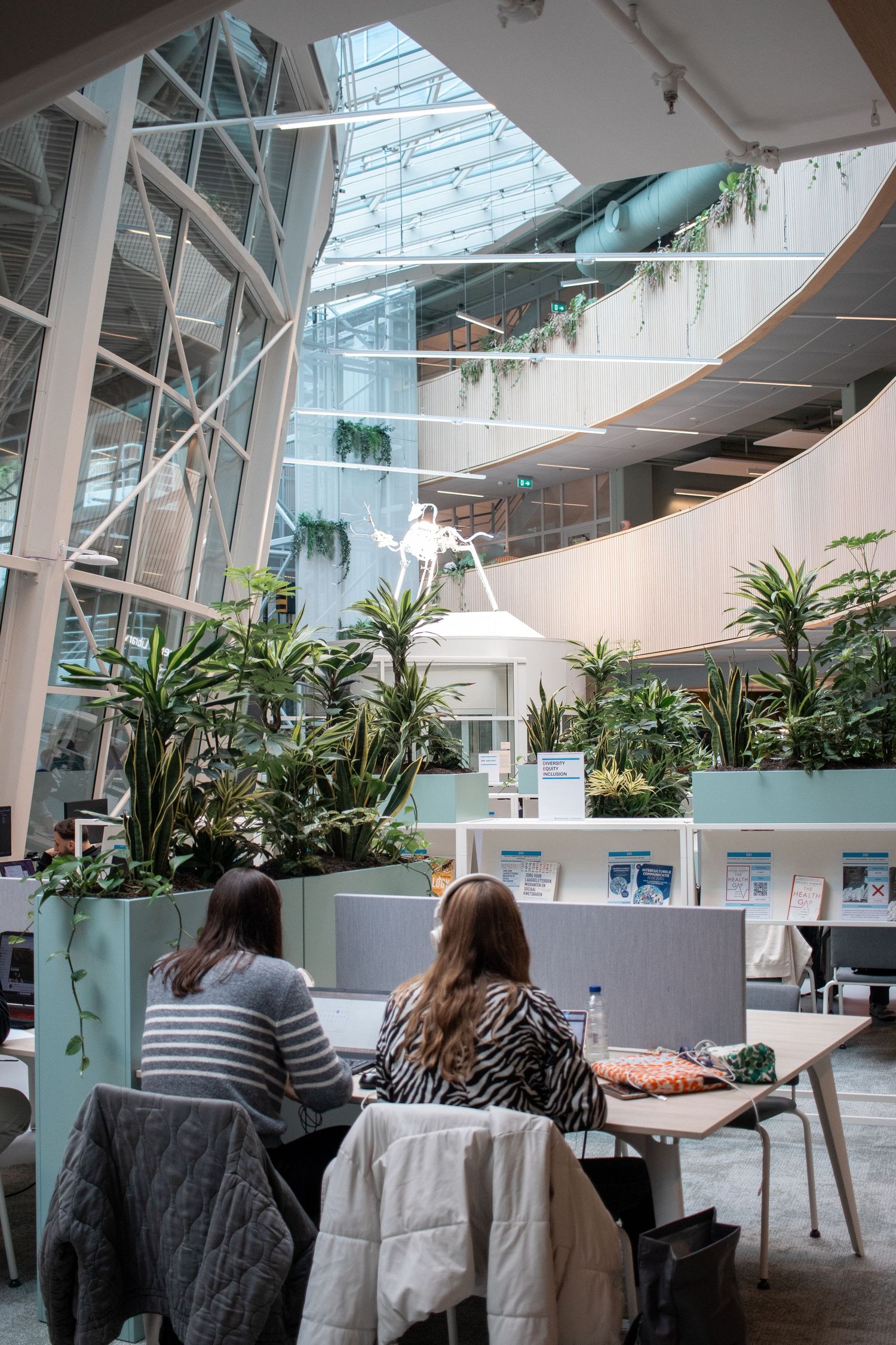 Two women studying at a desk in a modern atrium surrounded by lush tropical plants and glass ceiling.