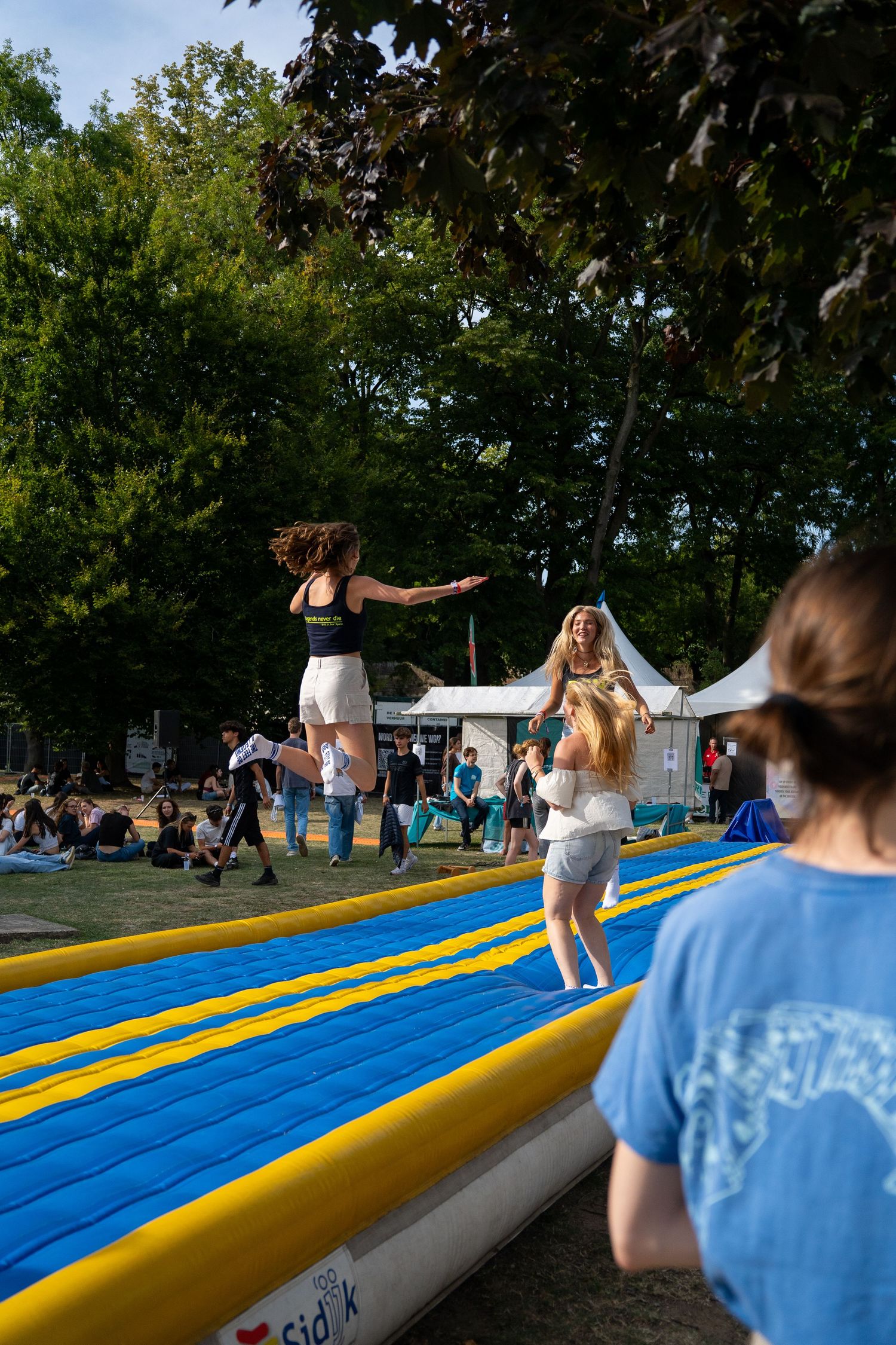 Two girls jumping on a blue and yellow inflatable air track at an outdoor summer festival.
