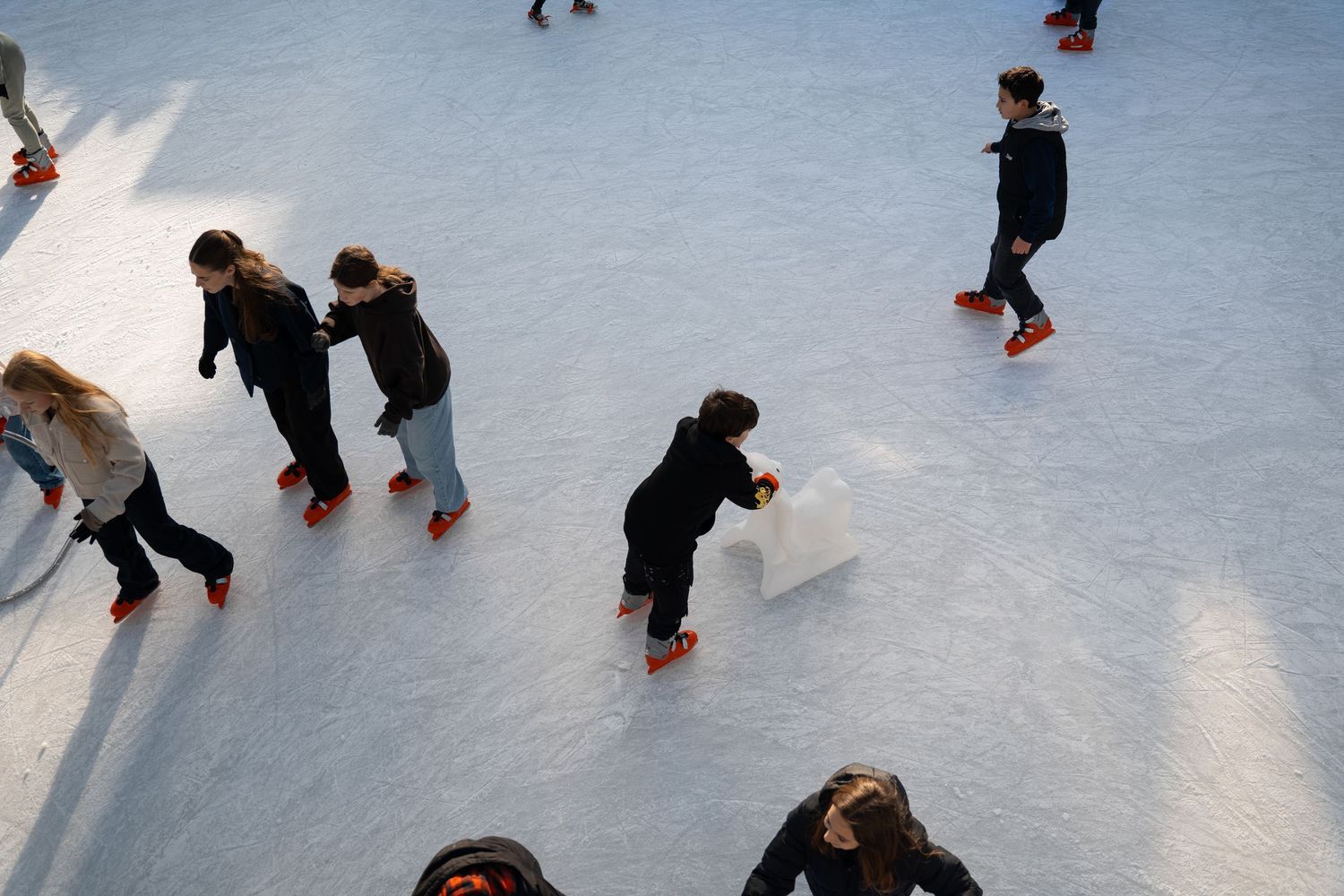 Overhead view of children and adults ice skating with orange-bladed skates on a white rink.