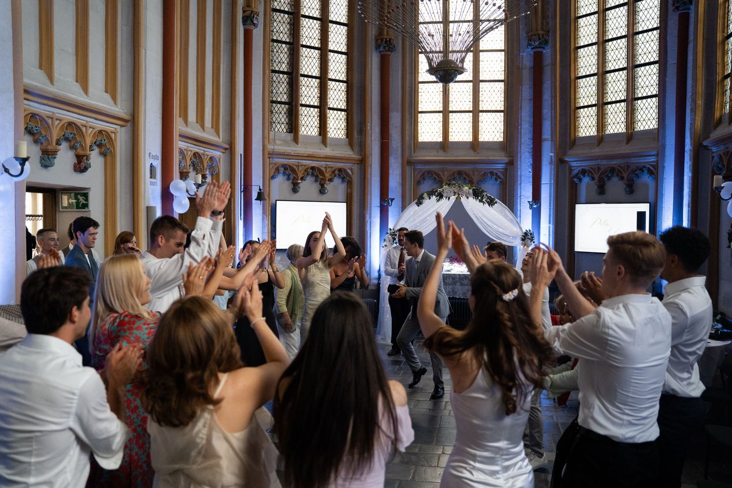 Wedding guests raise their hands and celebrate on the dance floor inside a grand ornate ballroom.