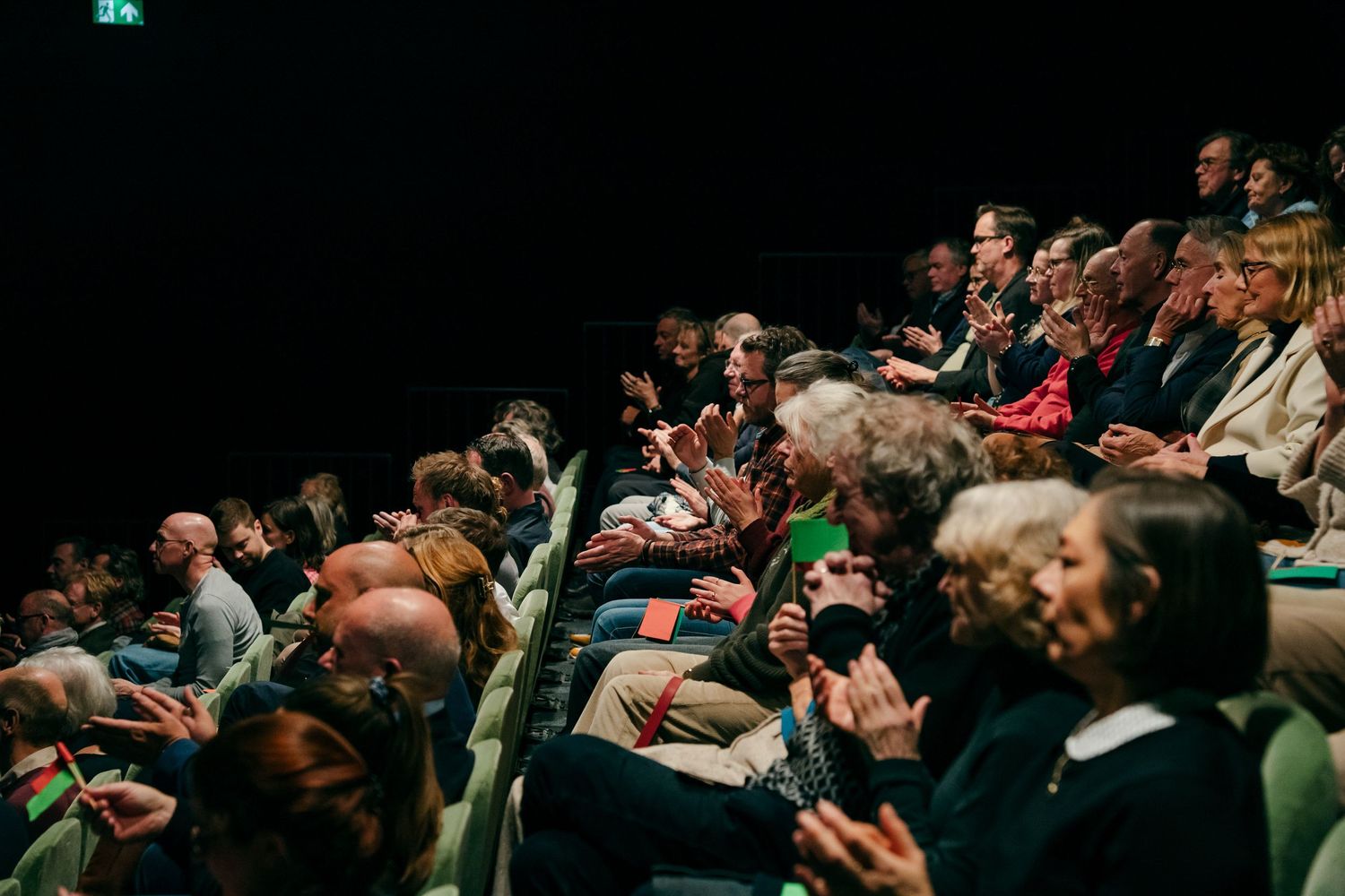 Rows of engaged audience members clapping in a green-seated theater during a live show.