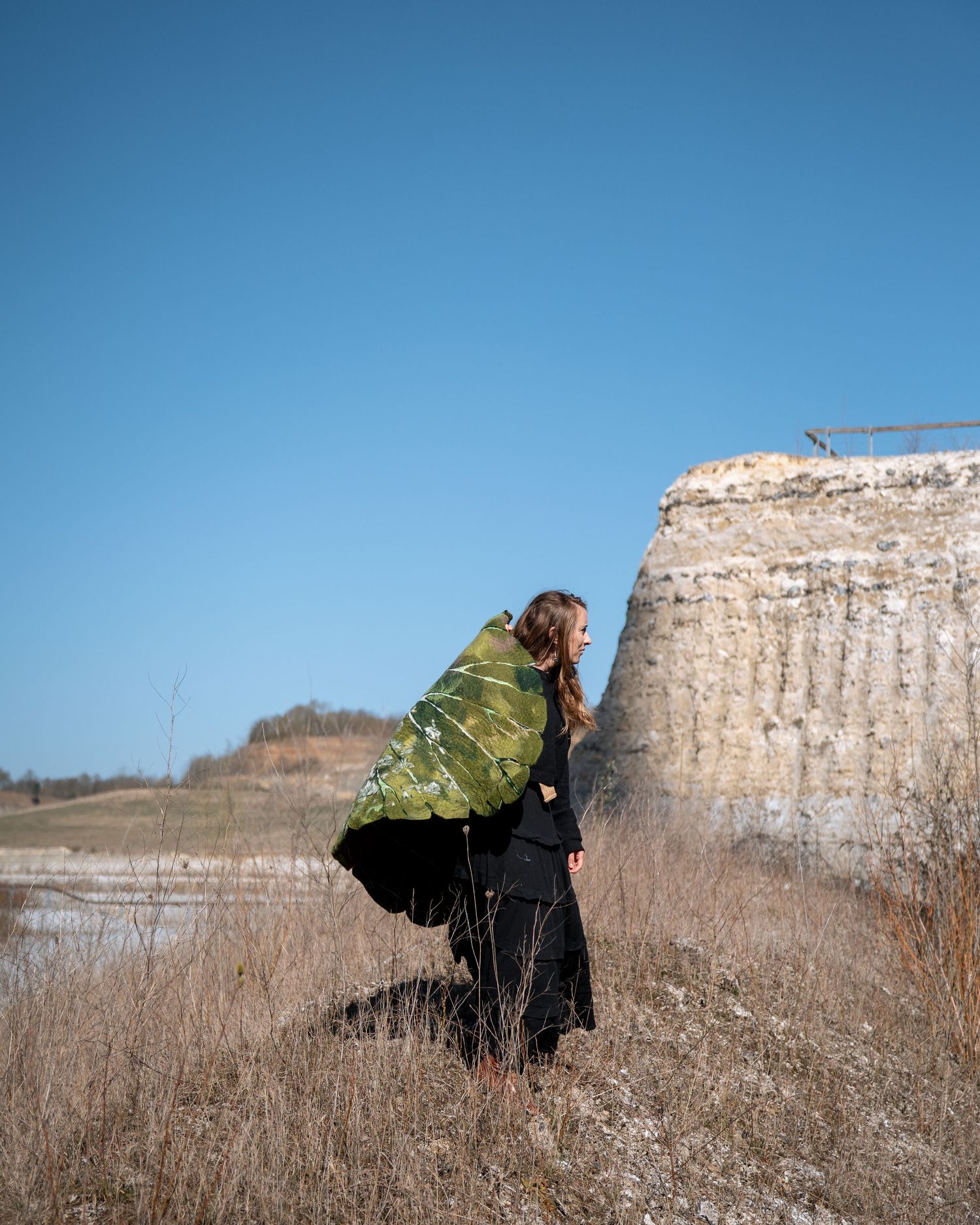 Woman in black dress wrapped in green textile standing beside a rocky cliff under blue sky.