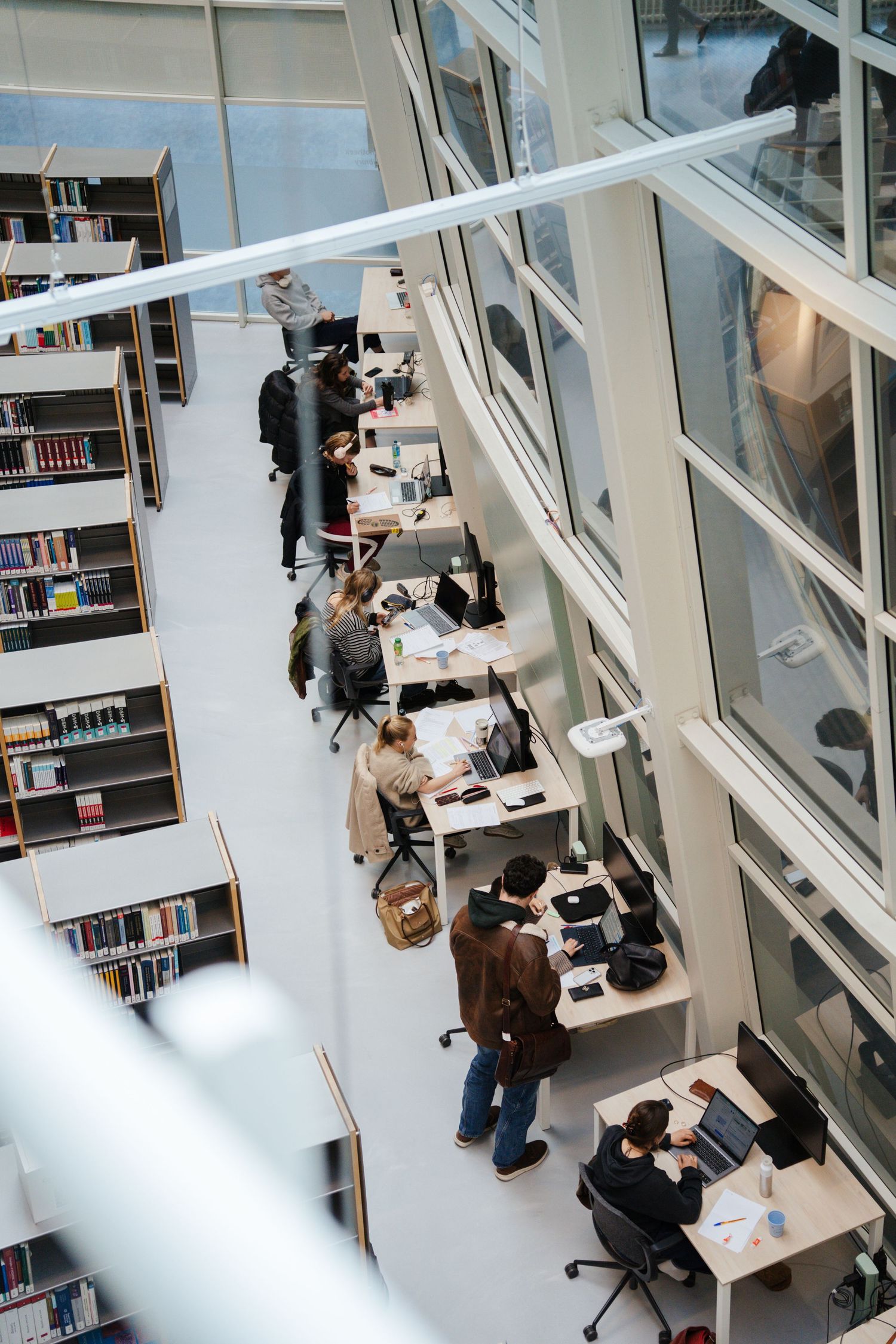 Students study and work on laptops at desks in a modern university library with floor-to-ceiling windows.