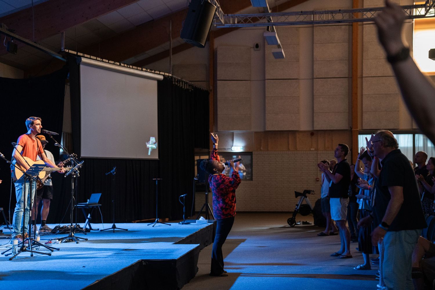 Guitarist leads worship on stage as attendees raise hands during a Christian praise event in a large hall.