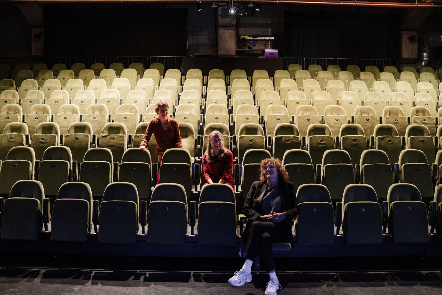 Three people sitting among rows of empty yellow theater seats in a dimly lit auditorium.