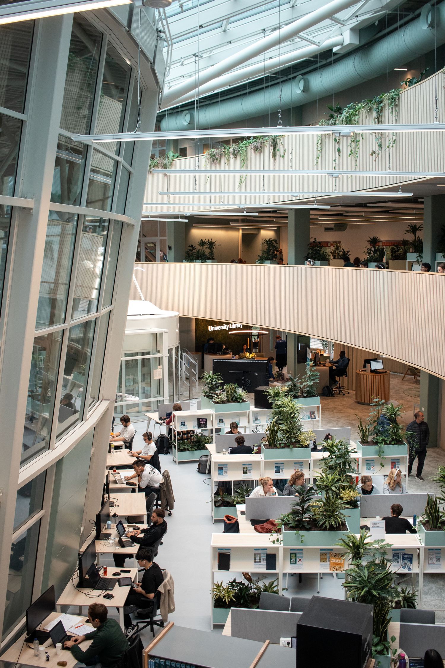 Busy modern open-plan university workspace with multiple floors, plants, and glass walls flooded with light.